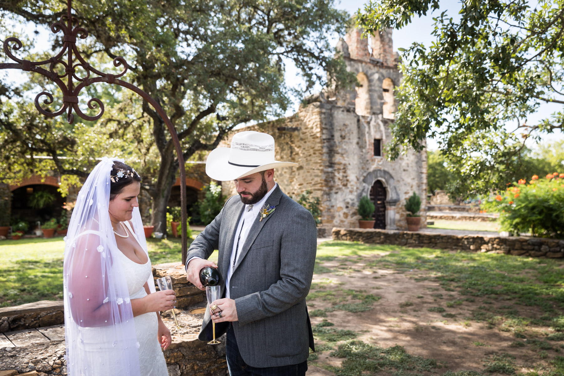 Bride in veil and groom wearing grey jacket and cowboy hat pouring champagne in front of church