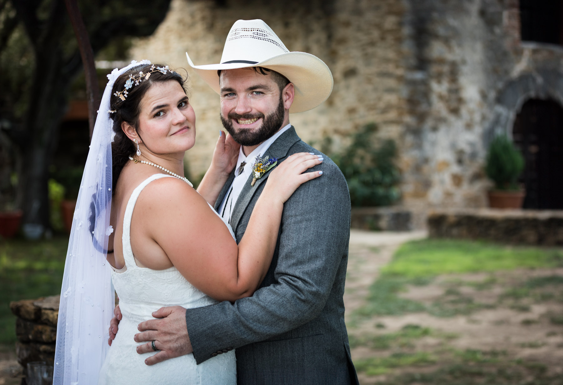 Bride in white sleeveless dress and groom wearing grey jacket and cowboy hat hugging in front of church for an article on how to get married at Mission Espada