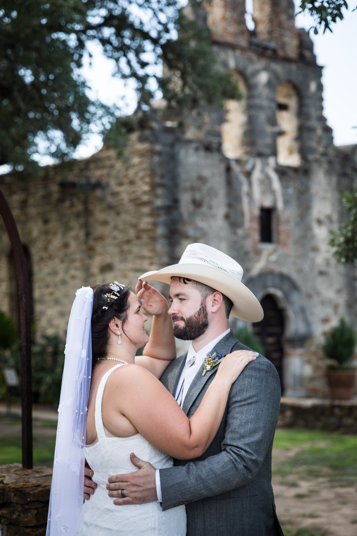 Bride in white sleeveless dress and groom wearing grey jacket and cowboy hat hugging in front of church for an article on how to get married at Mission Espada