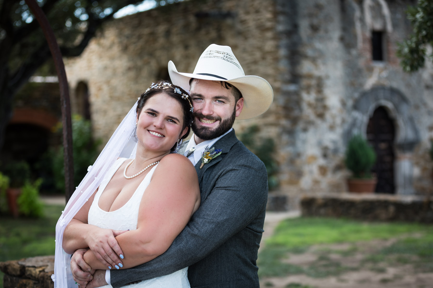 Bride in white sleeveless dress and groom wearing grey jacket and cowboy hat hugging in front of church for an article on how to get married at Mission Espada