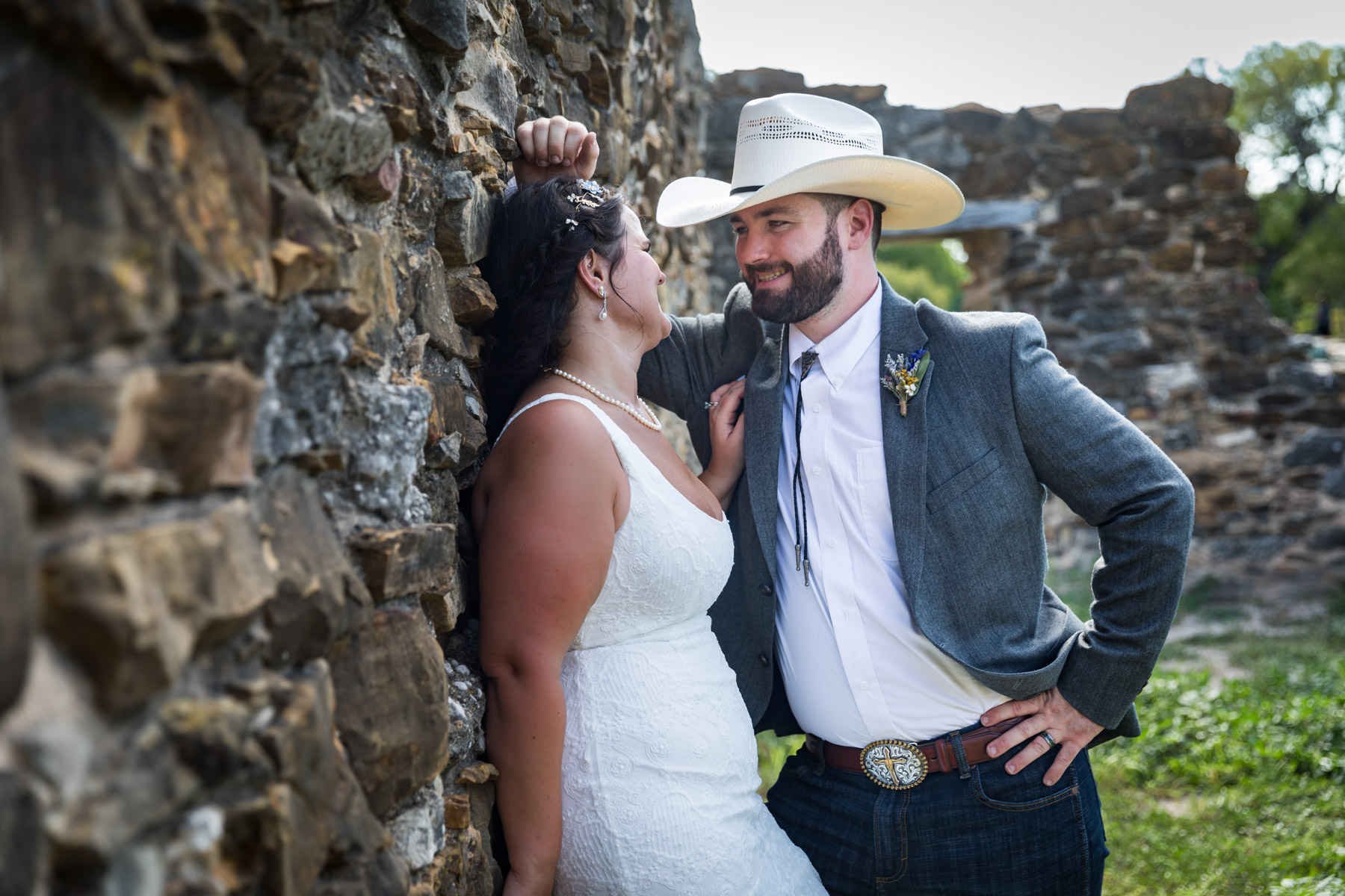 Bride in white sleeveless dress and groom wearing grey jacket and cowboy hat leaning against stone wall