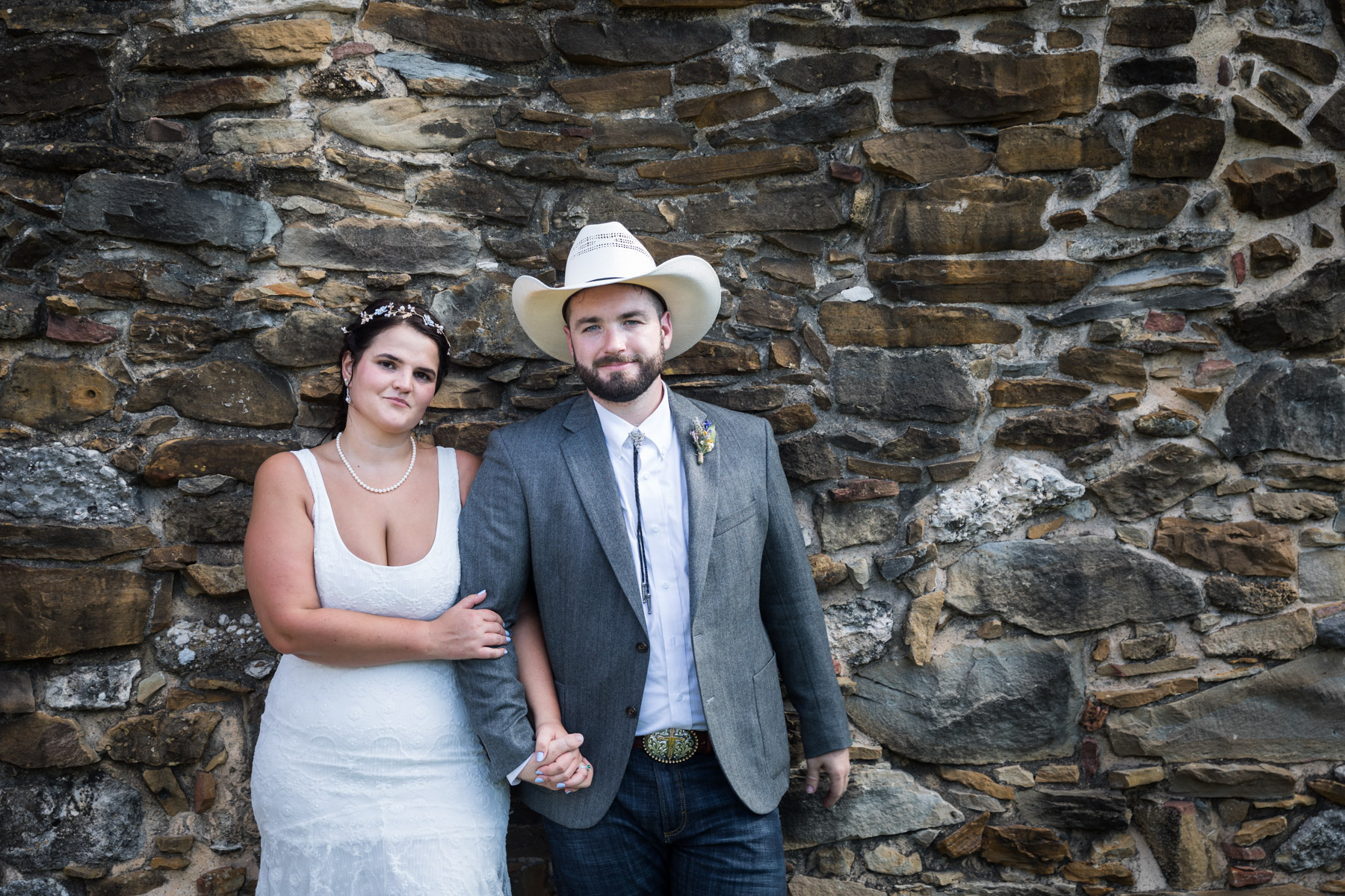 Bride in white sleeveless dress and groom wearing grey jacket and cowboy hat holding hands against stone wall