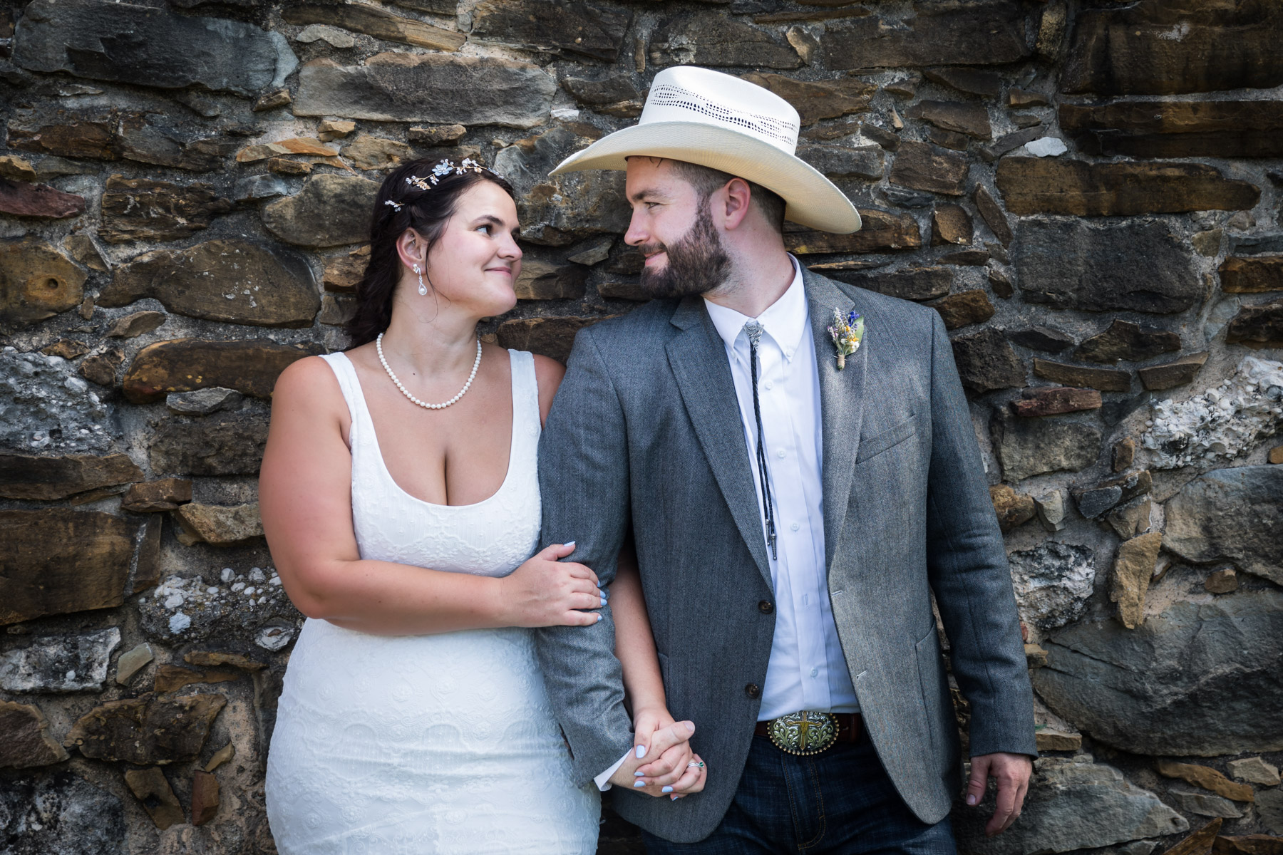 Bride in white sleeveless dress and groom wearing grey jacket and cowboy hat holding hands against stone wall