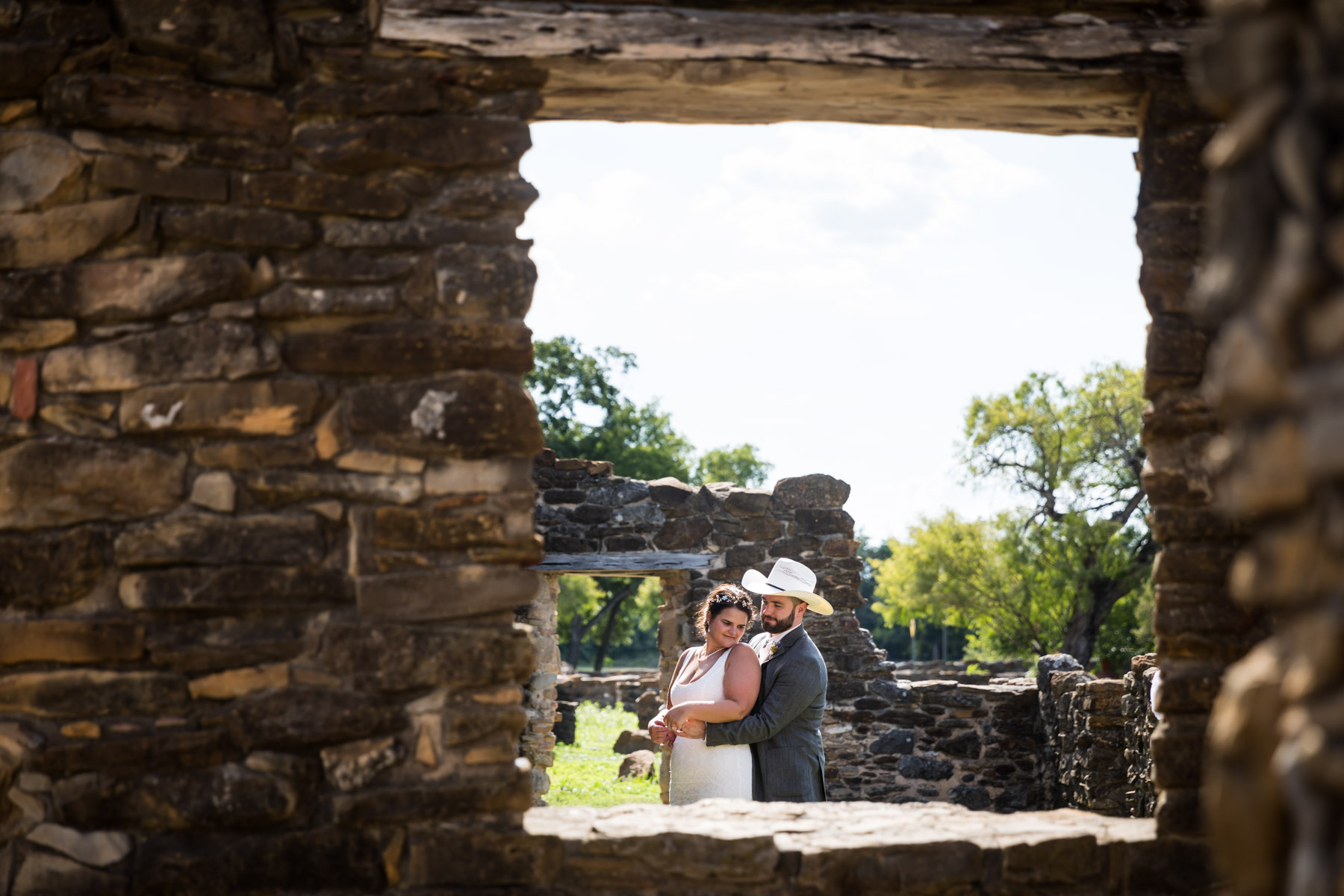 Bride in white sleeveless dress and groom wearing grey jacket and cowboy hat hugging in stone ruins for an article on how to get married at Mission Espada