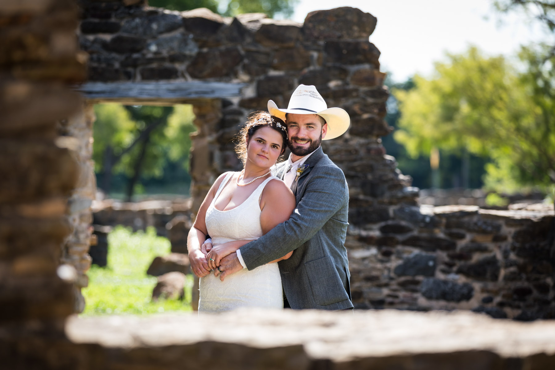 Bride in white sleeveless dress and groom wearing grey jacket and cowboy hat hugging in stone ruins for an article on how to get married at Mission Espada