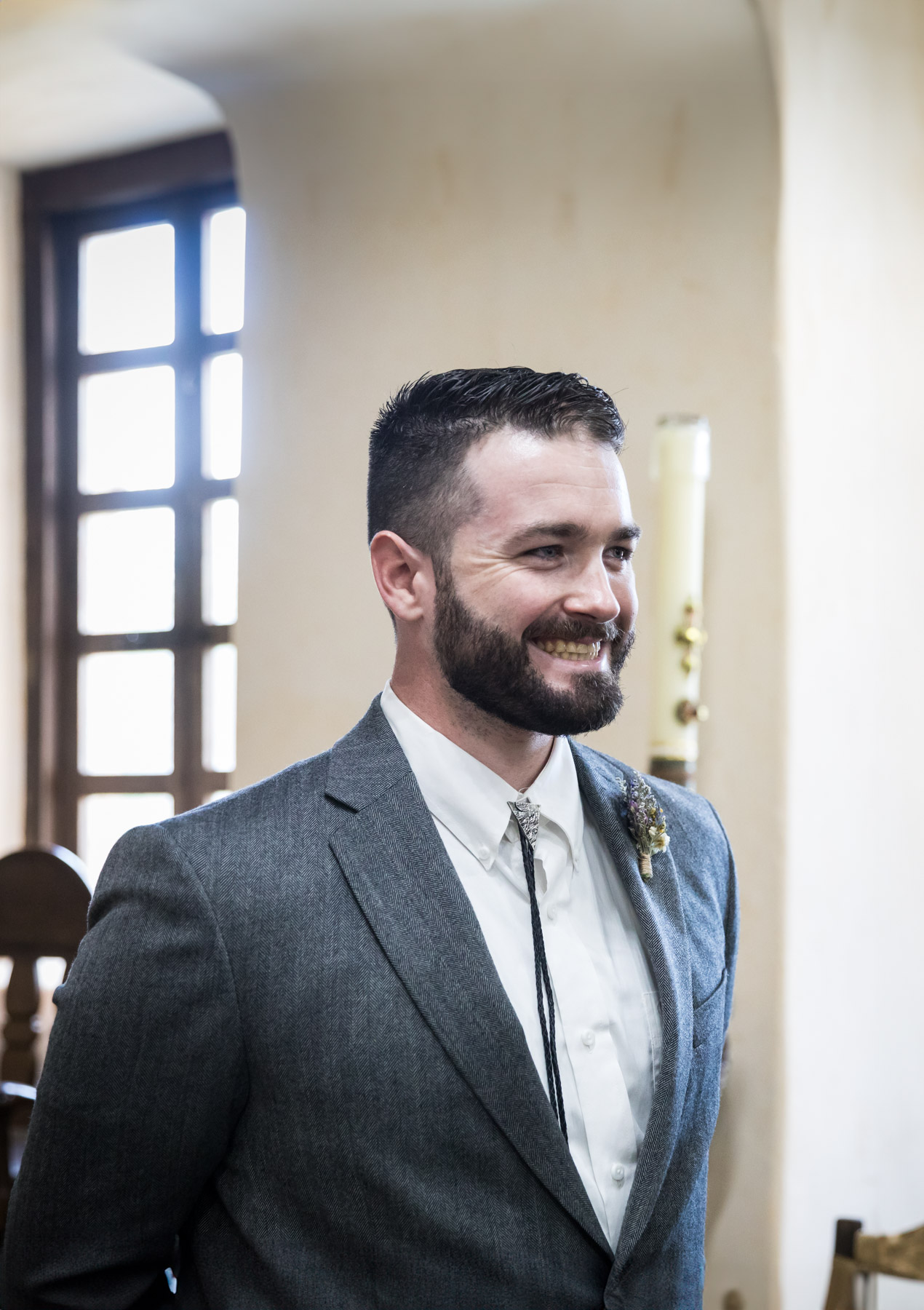 Groom wearing bolo tie and grey jacket smiling for an article on how to get married at Mission Espada