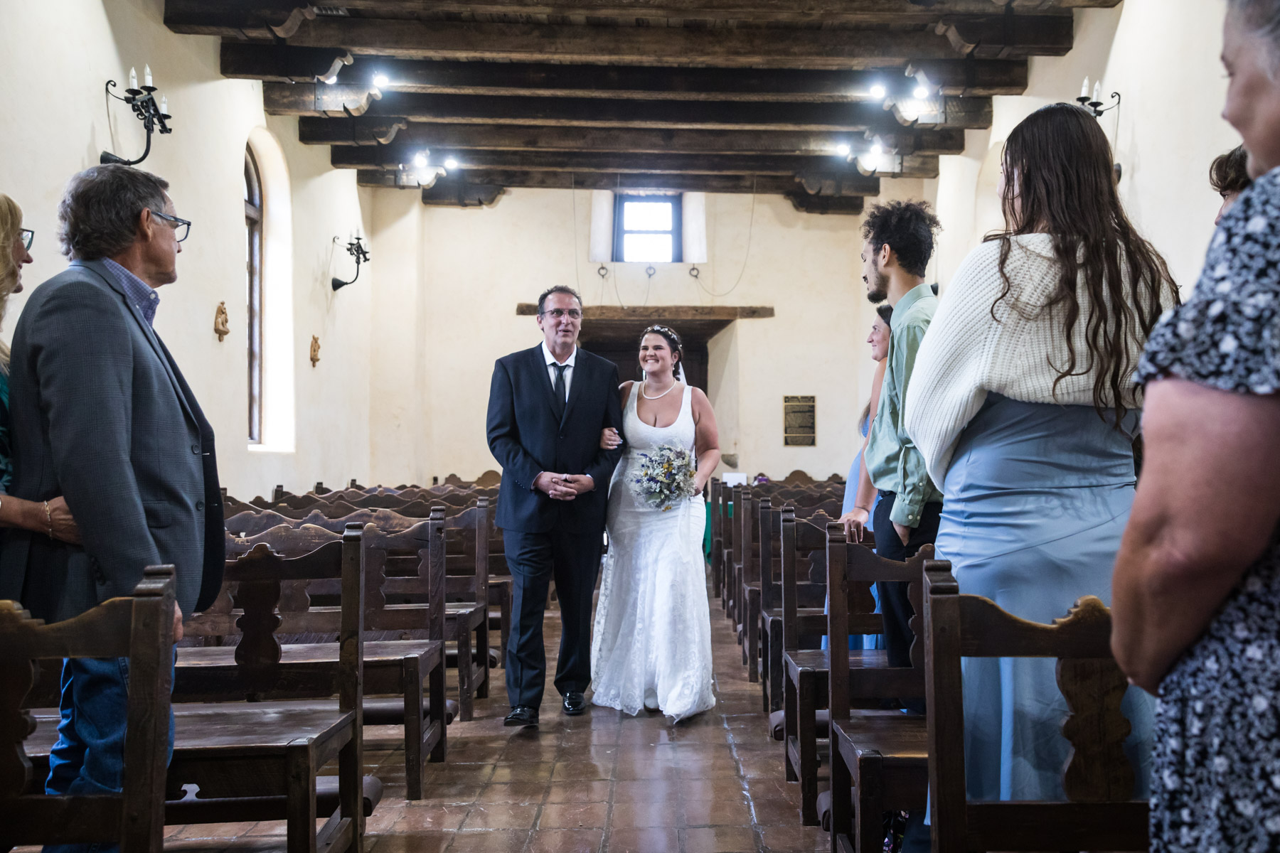 Bride and father walking up aisle towards guests during ceremony for an article on how to get married at Mission Espada