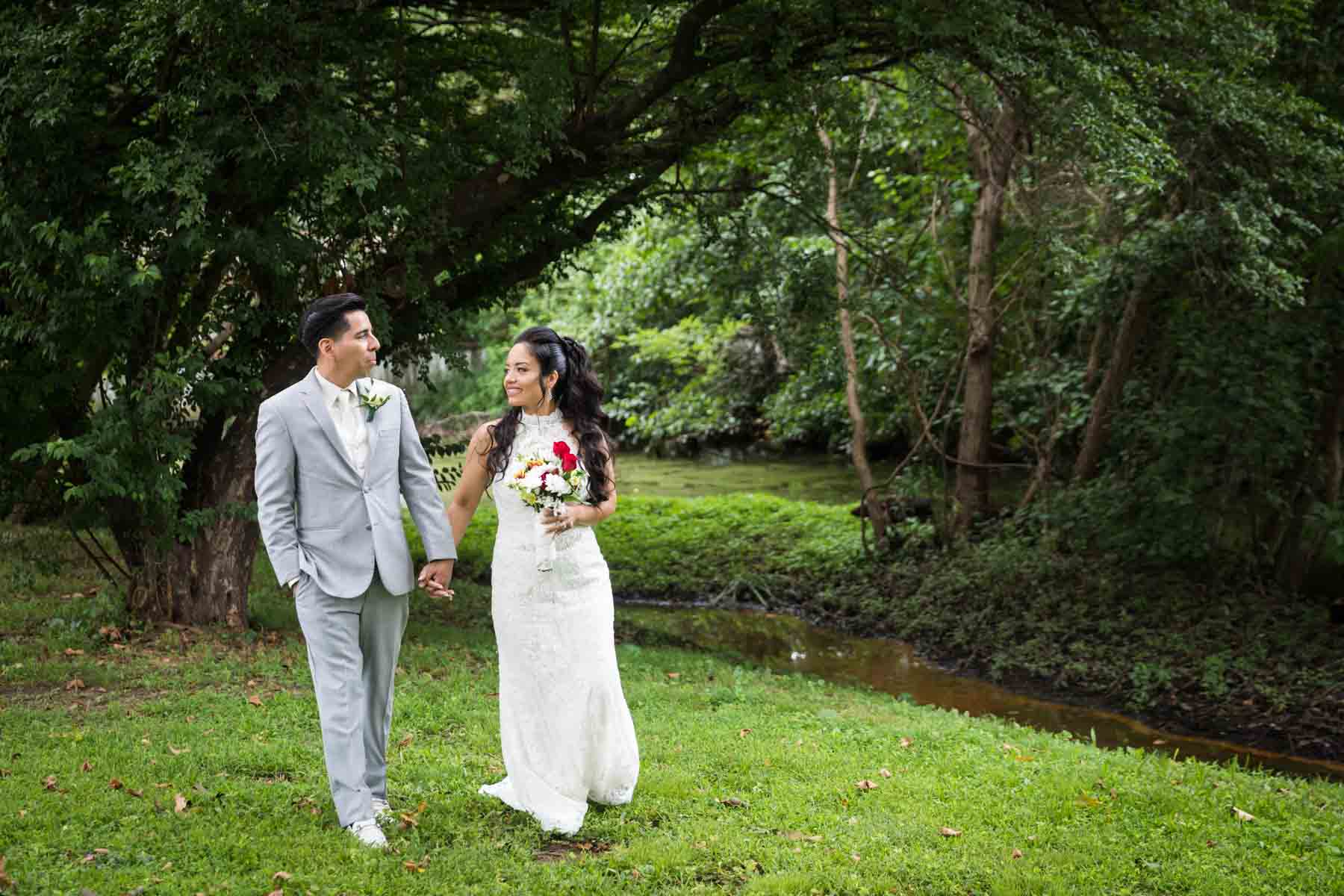 Bride and groom holding hands and standing in grass in front of creek and trees for an article on how to have an eco-friendly wedding