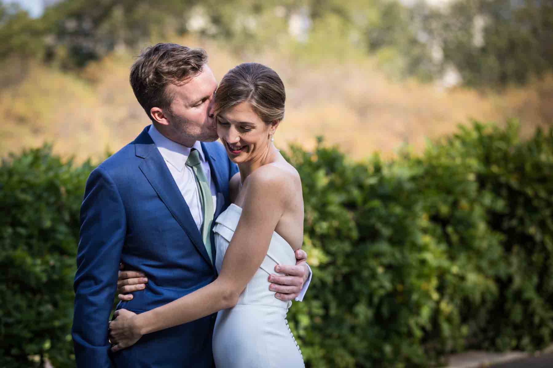 Groom kissing bride on the side of head in front of plants for an article on how to have an eco-friendly wedding