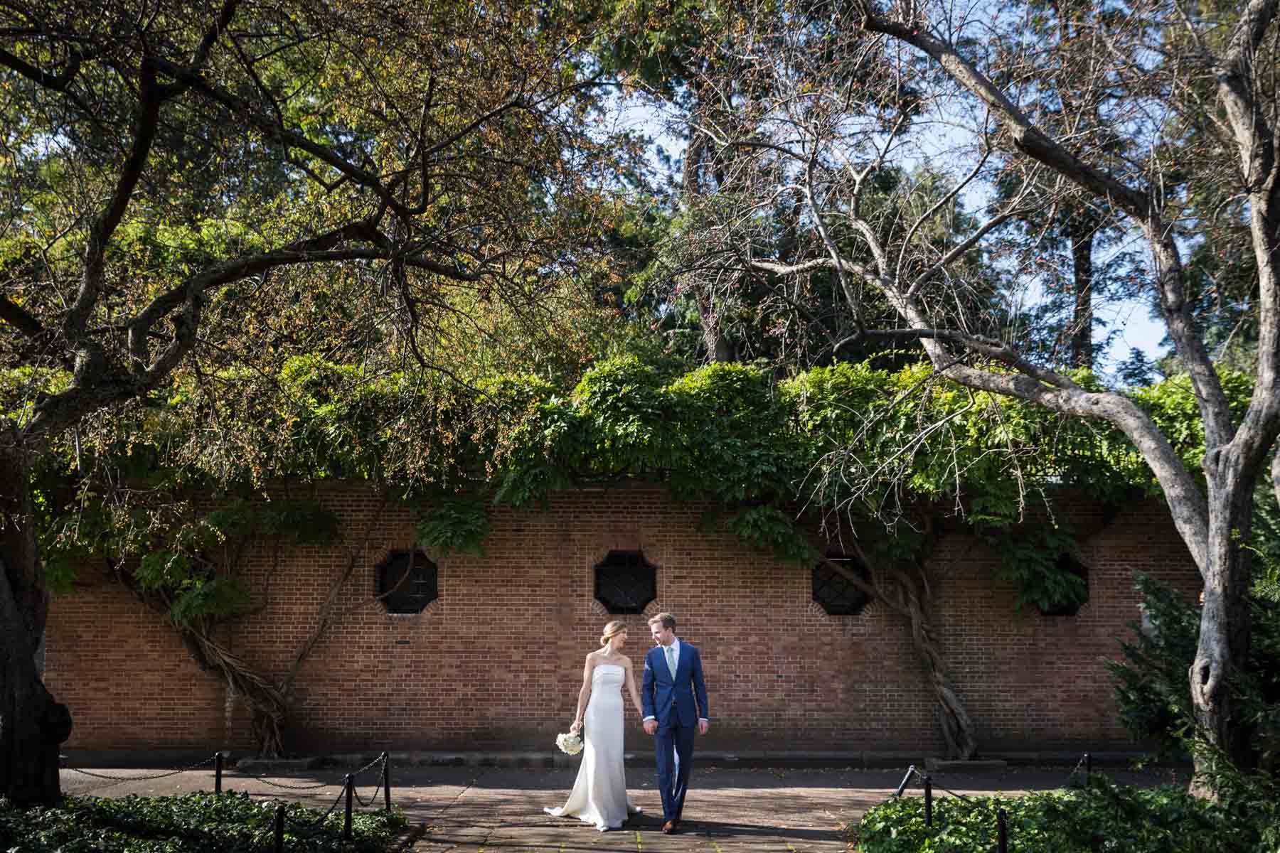 Bride and groom holding hands walking down pathway in front of brick wall and vines for an article on how to have an eco-friendly wedding