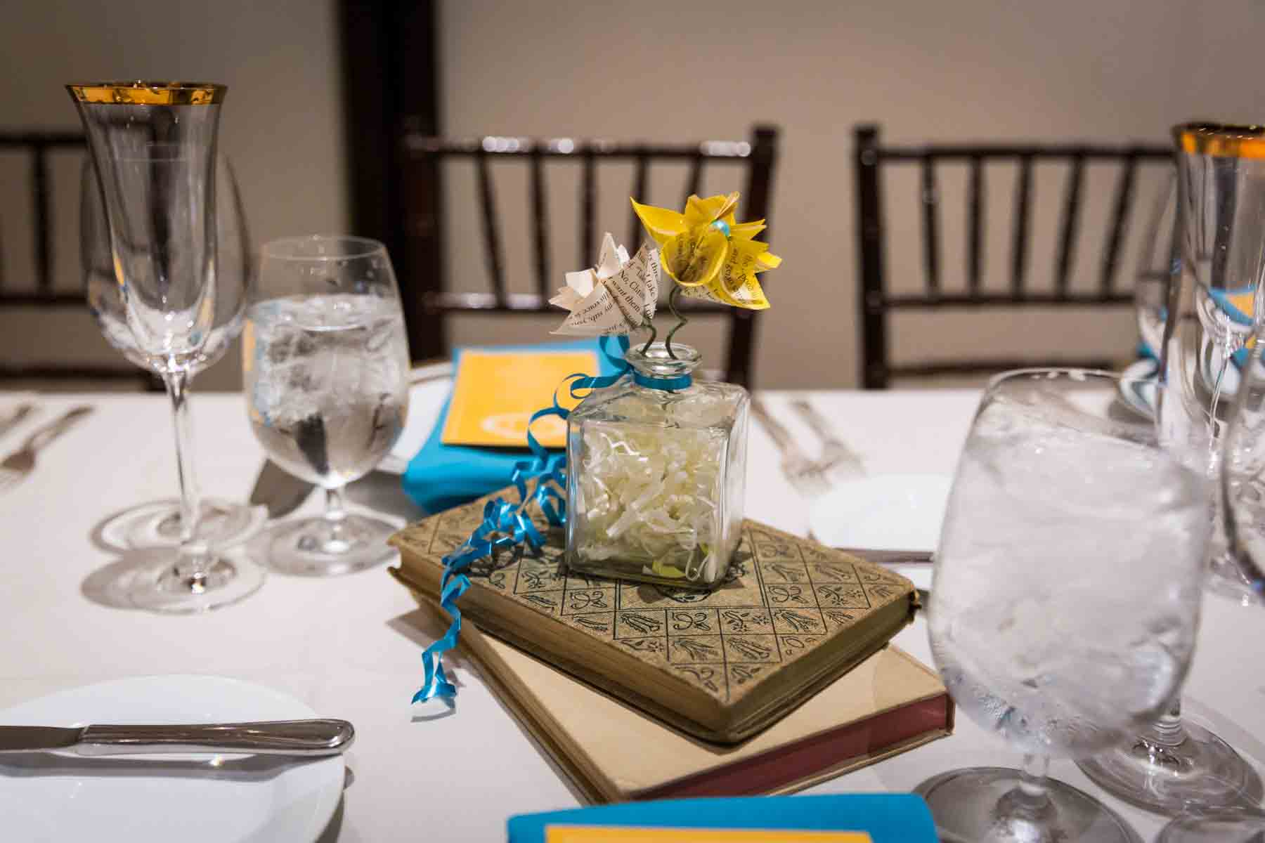 Table centerpiece made of books and glass vase with paper flowers and blue ribbons