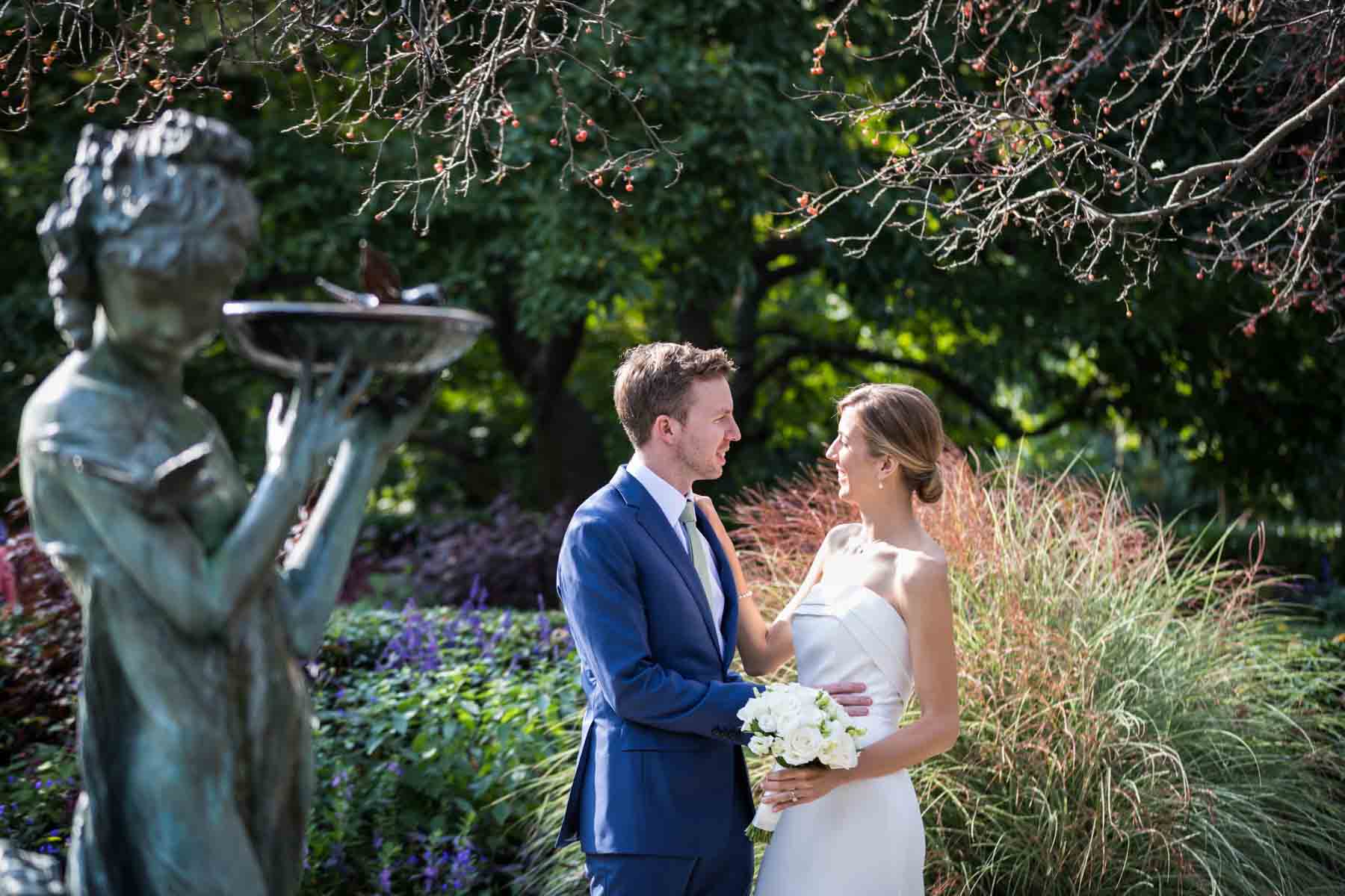 Bride and groom looking at each other with bronze statue to the side and reeds and purple flowers in the back