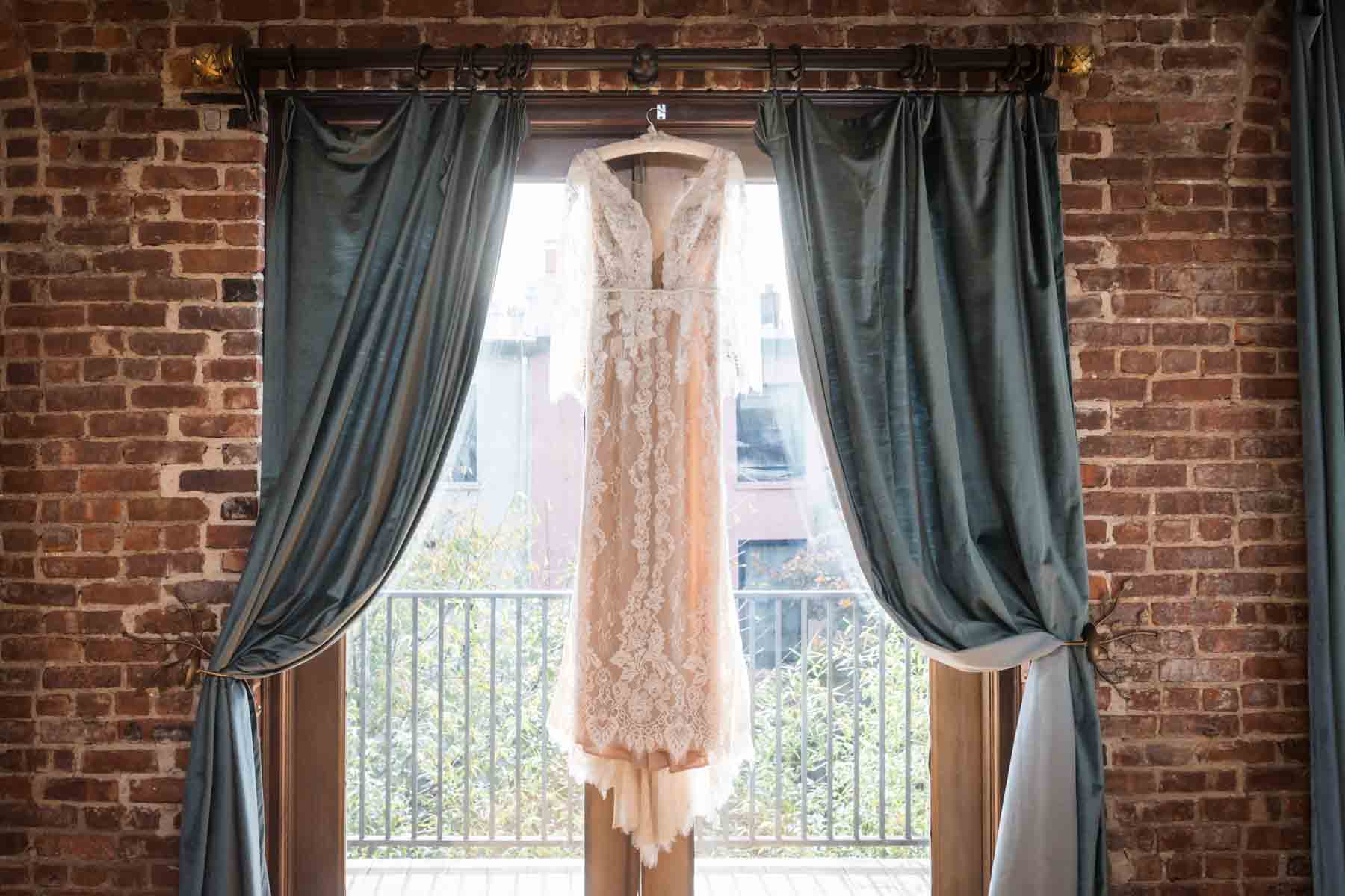 Close up on wedding dress hanging from curtain rod in front of window and brick wall