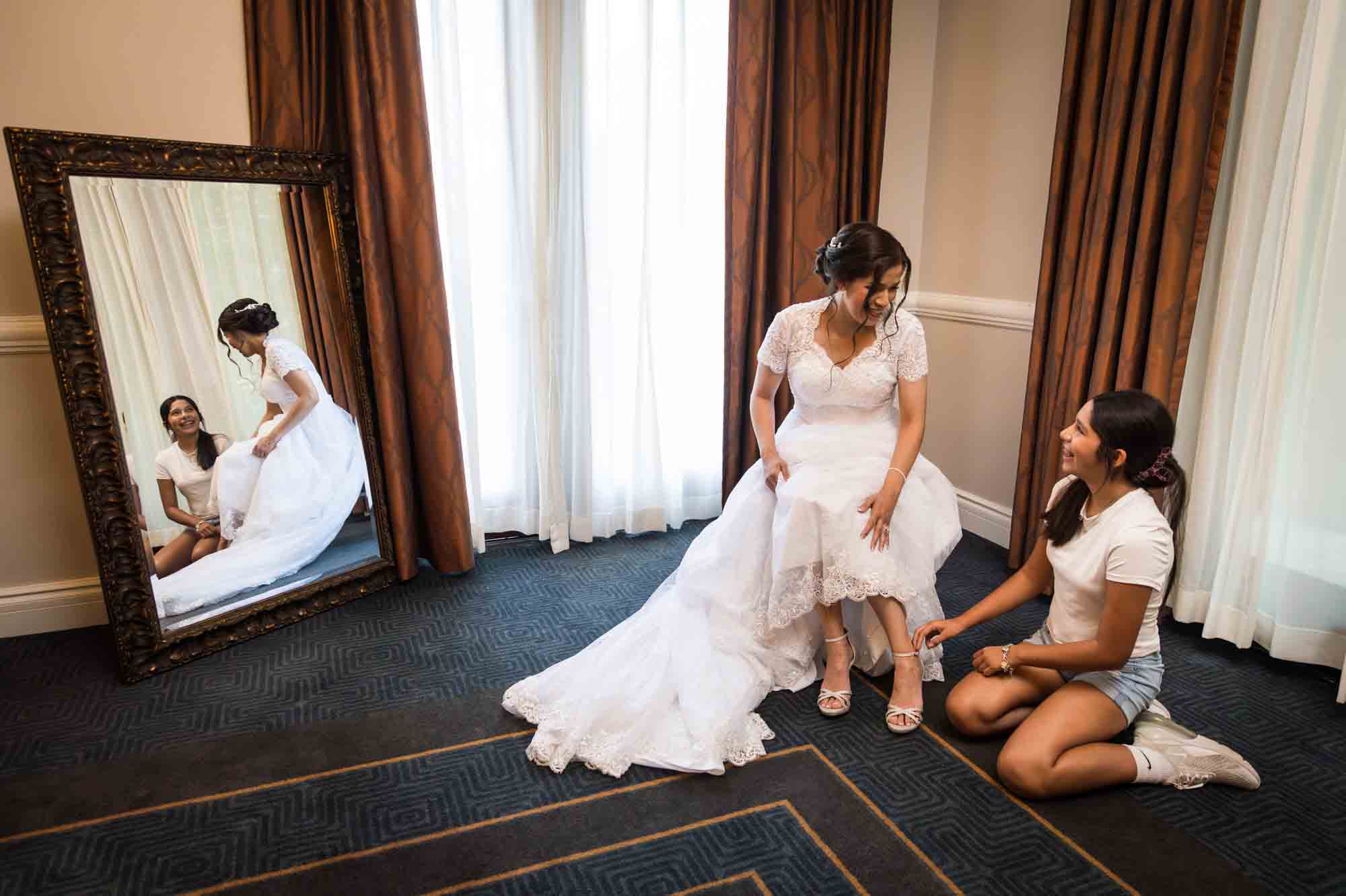 Bride seated with young girl helping her with shoes, reflected in mirror during a Hotel Contessa bridal portrait session in downtown San Antonio