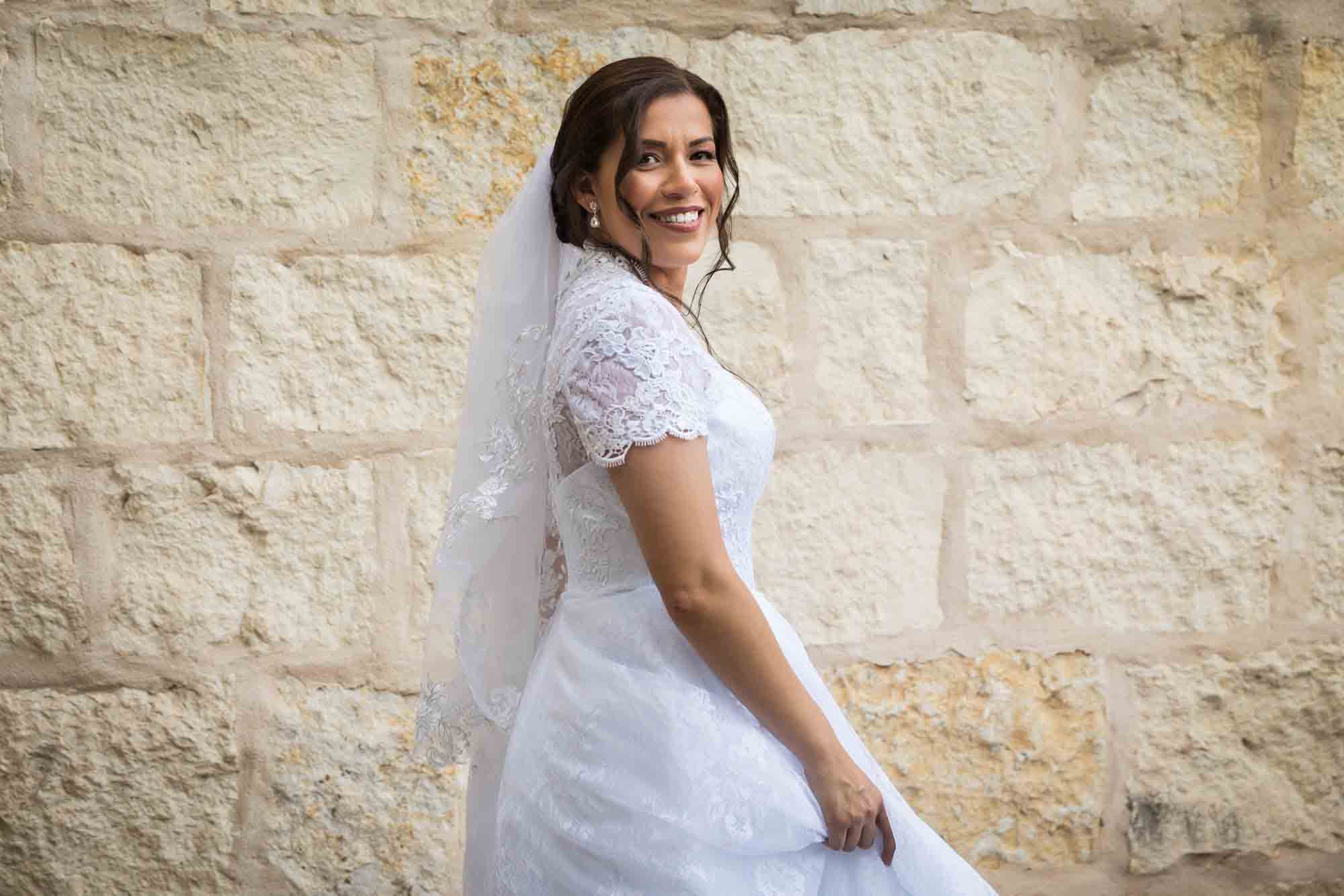 Bride posing in white dress with veil in front of beige, brick wall for an article about a downtown San Antonio bridal portrait session