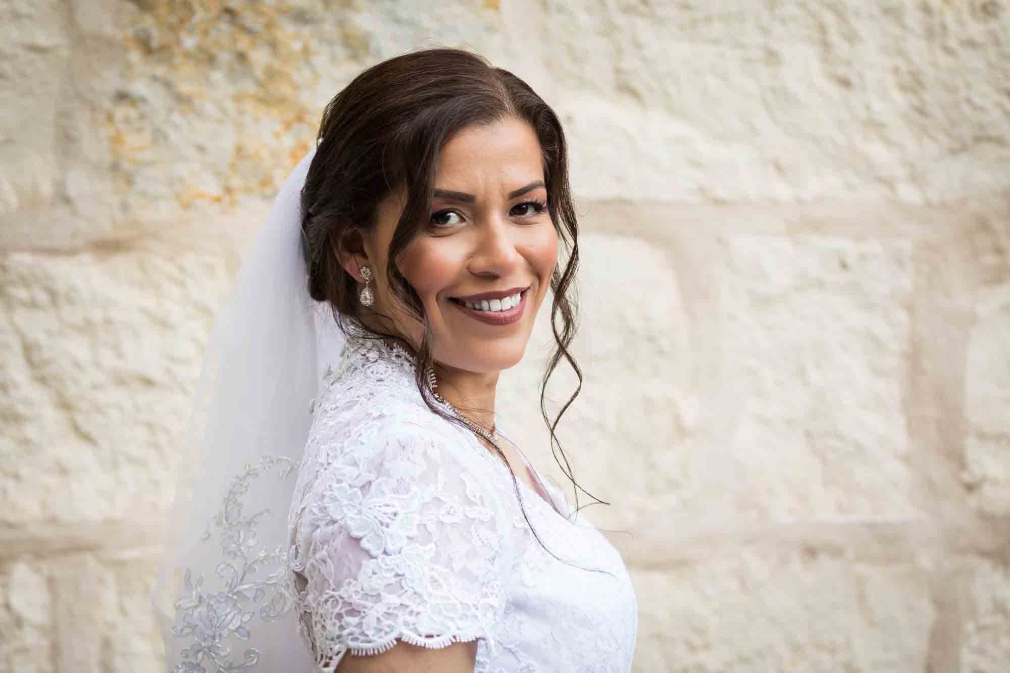 Bride posing in white dress with veil in front of beige, brick wall for an article about a downtown San Antonio bridal portrait session