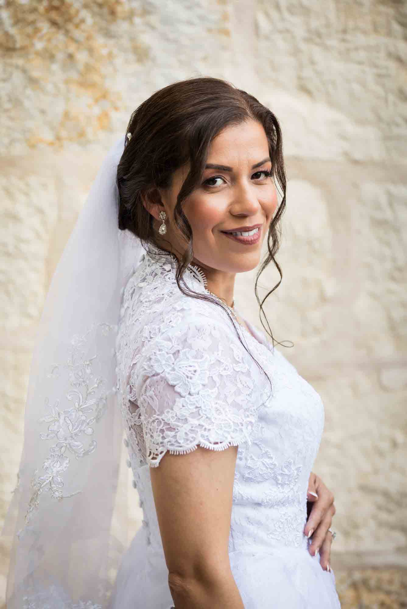 Bride posing in white dress with veil in front of beige, brick wall for an article about a downtown San Antonio bridal portrait session