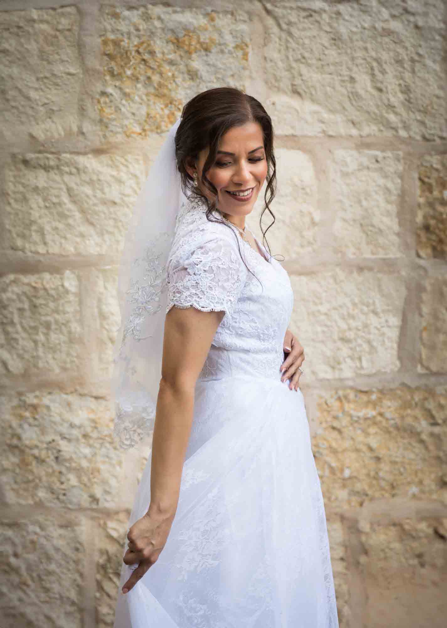 Bride posing in white dress with veil in front of beige, brick wall for an article about a downtown San Antonio bridal portrait session