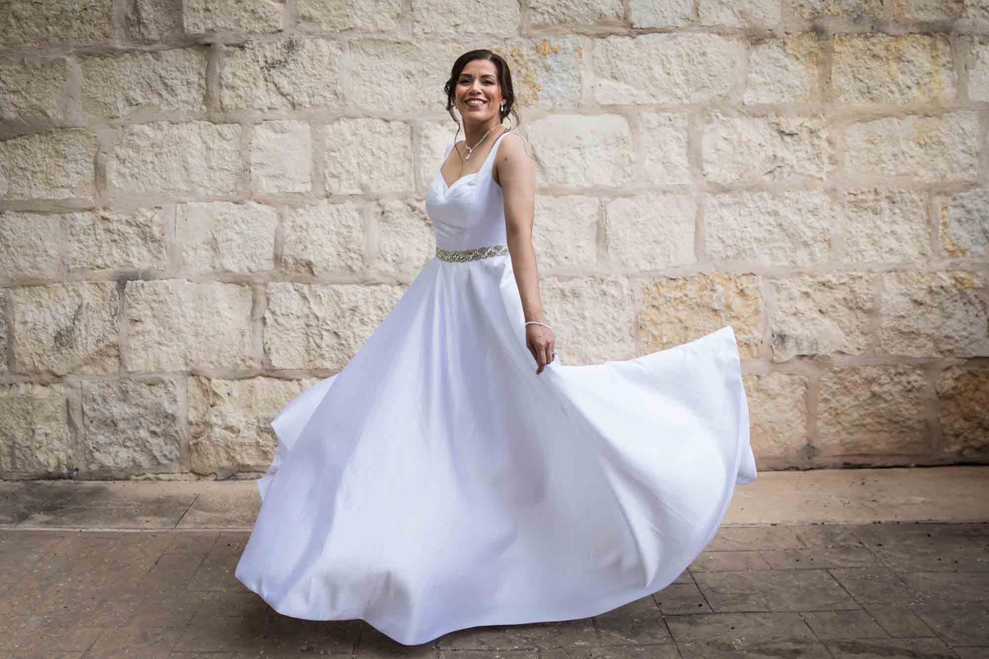 Bride twirling in white, sleeveless dress in front of beige, brick wall for an article about a downtown San Antonio bridal portrait session