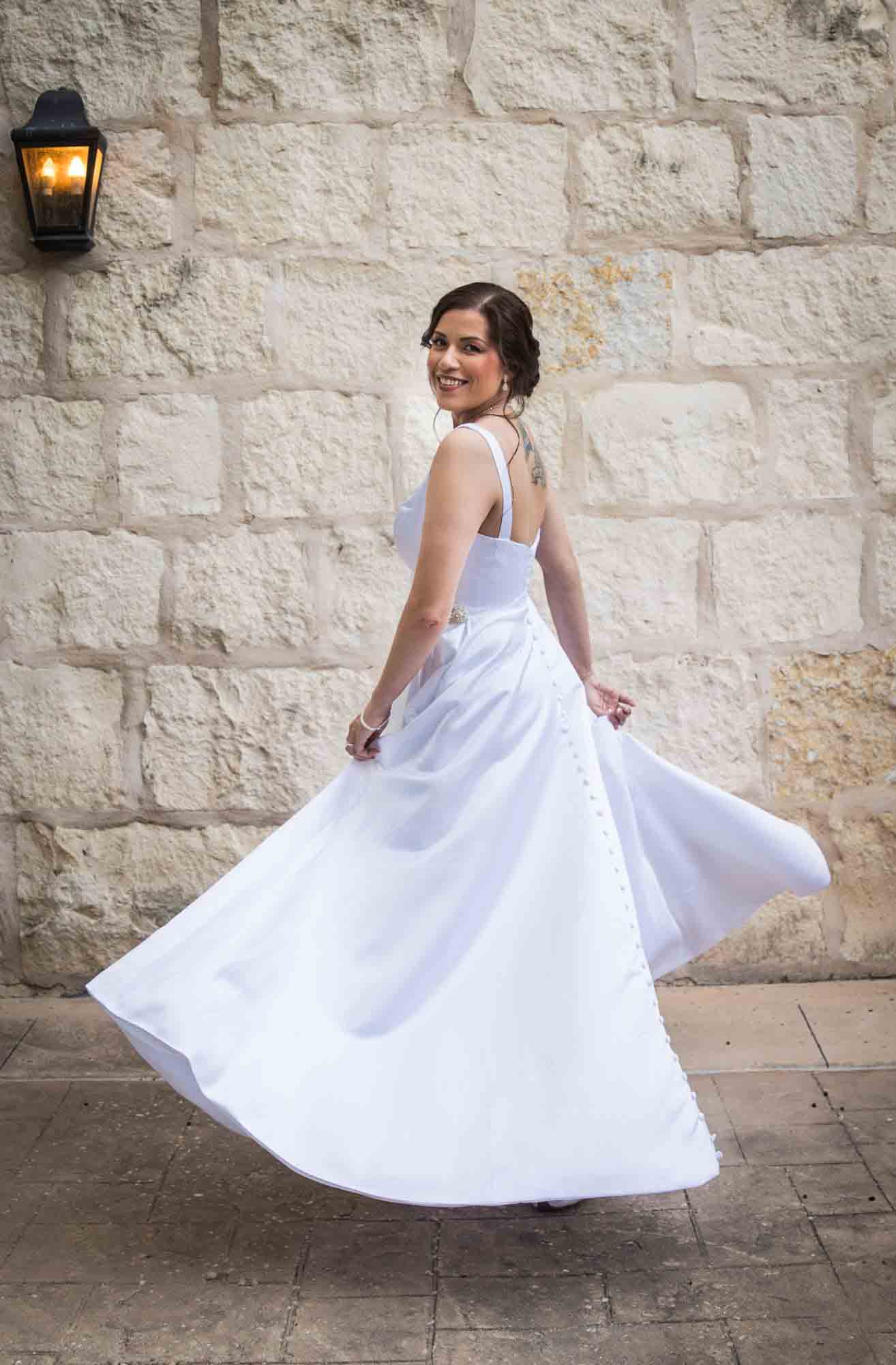 Bride twirling in white, sleeveless dress in front of beige, brick wall for an article about a downtown San Antonio bridal portrait session