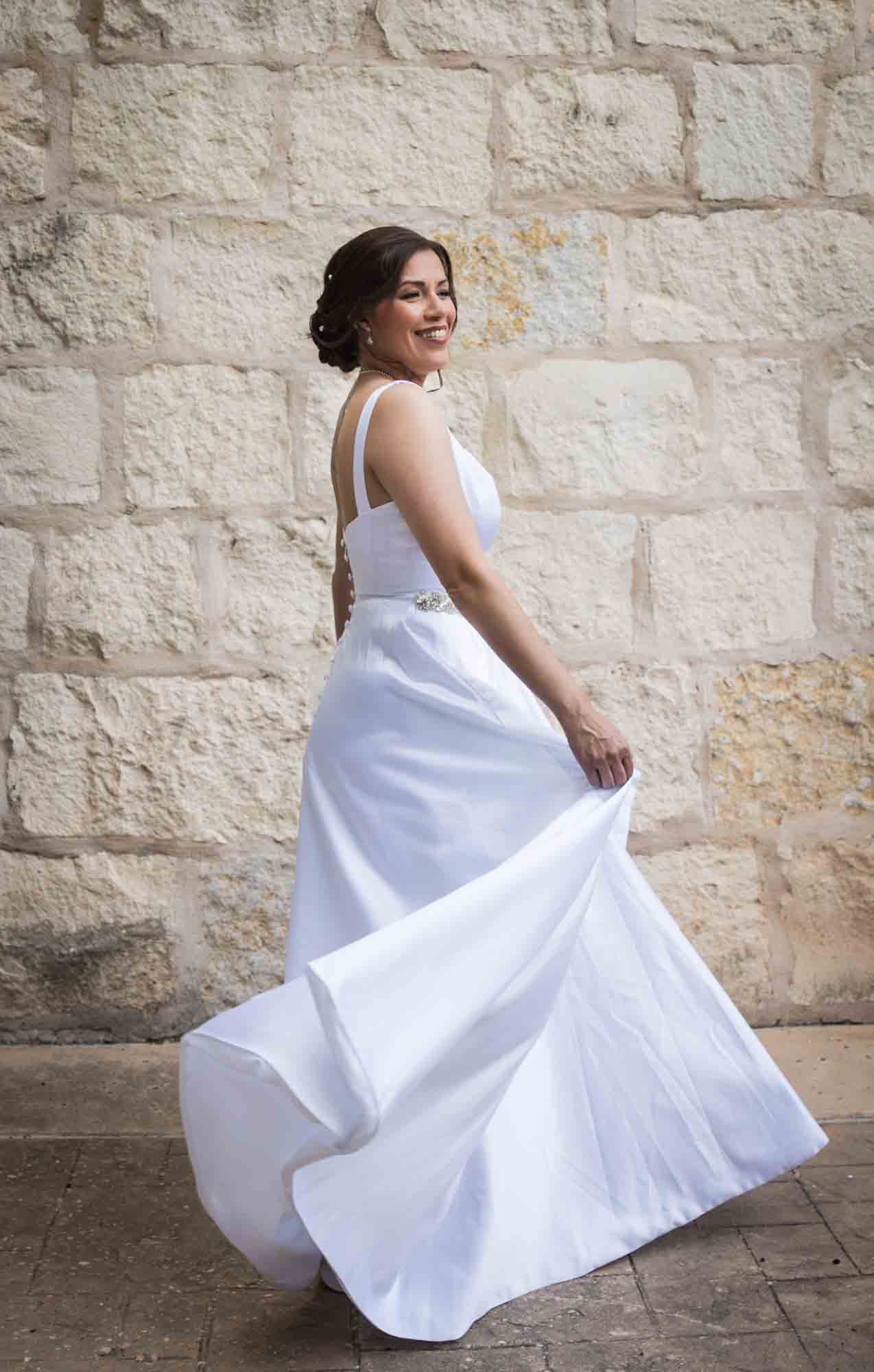 Bride twirling in white, sleeveless dress in front of beige, brick wall for an article about a downtown San Antonio bridal portrait session