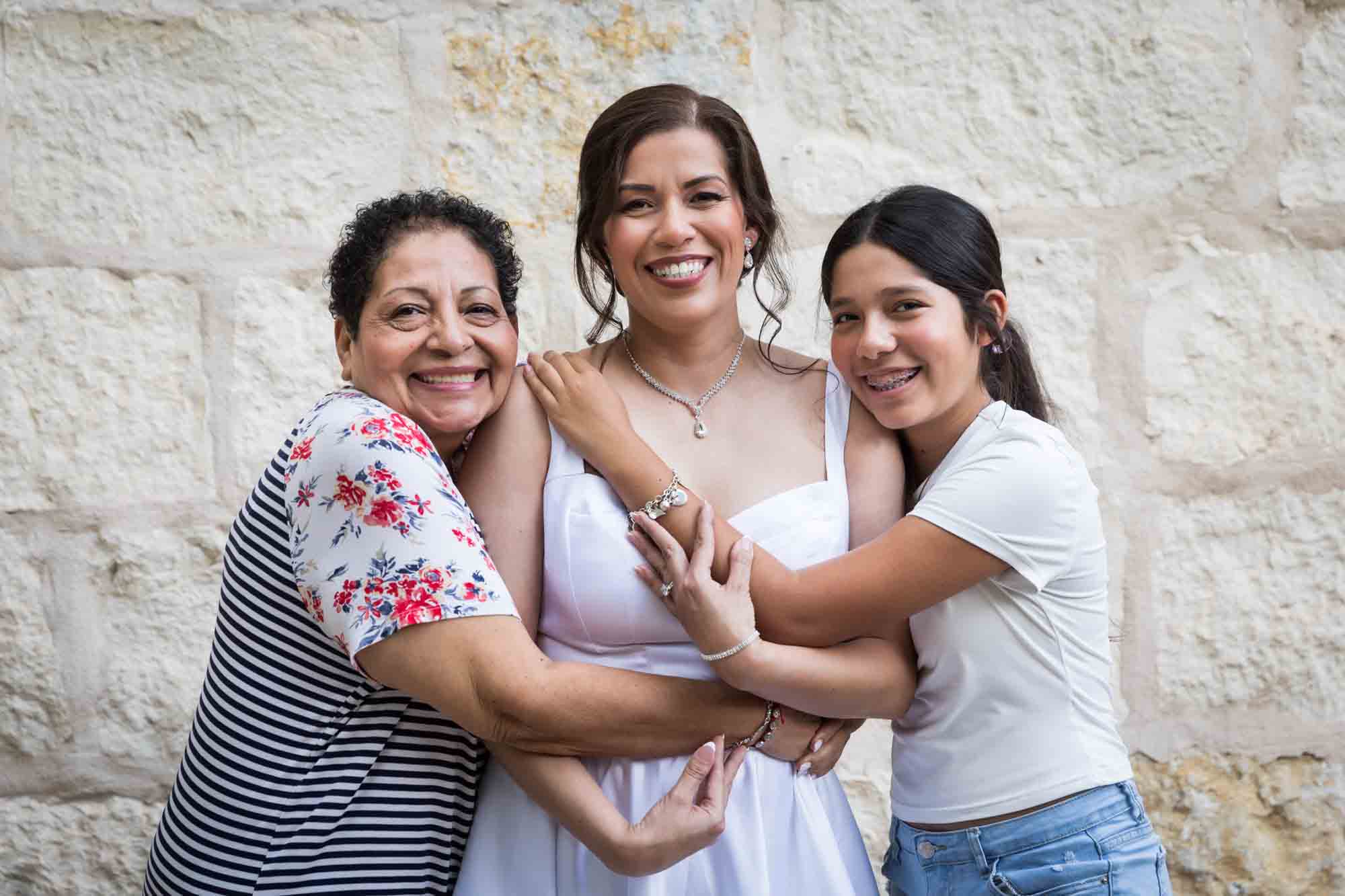 Bride, mother-in-law and young girl hugging in front of beige, brick wall during a downtown San Antonio bridal portrait session