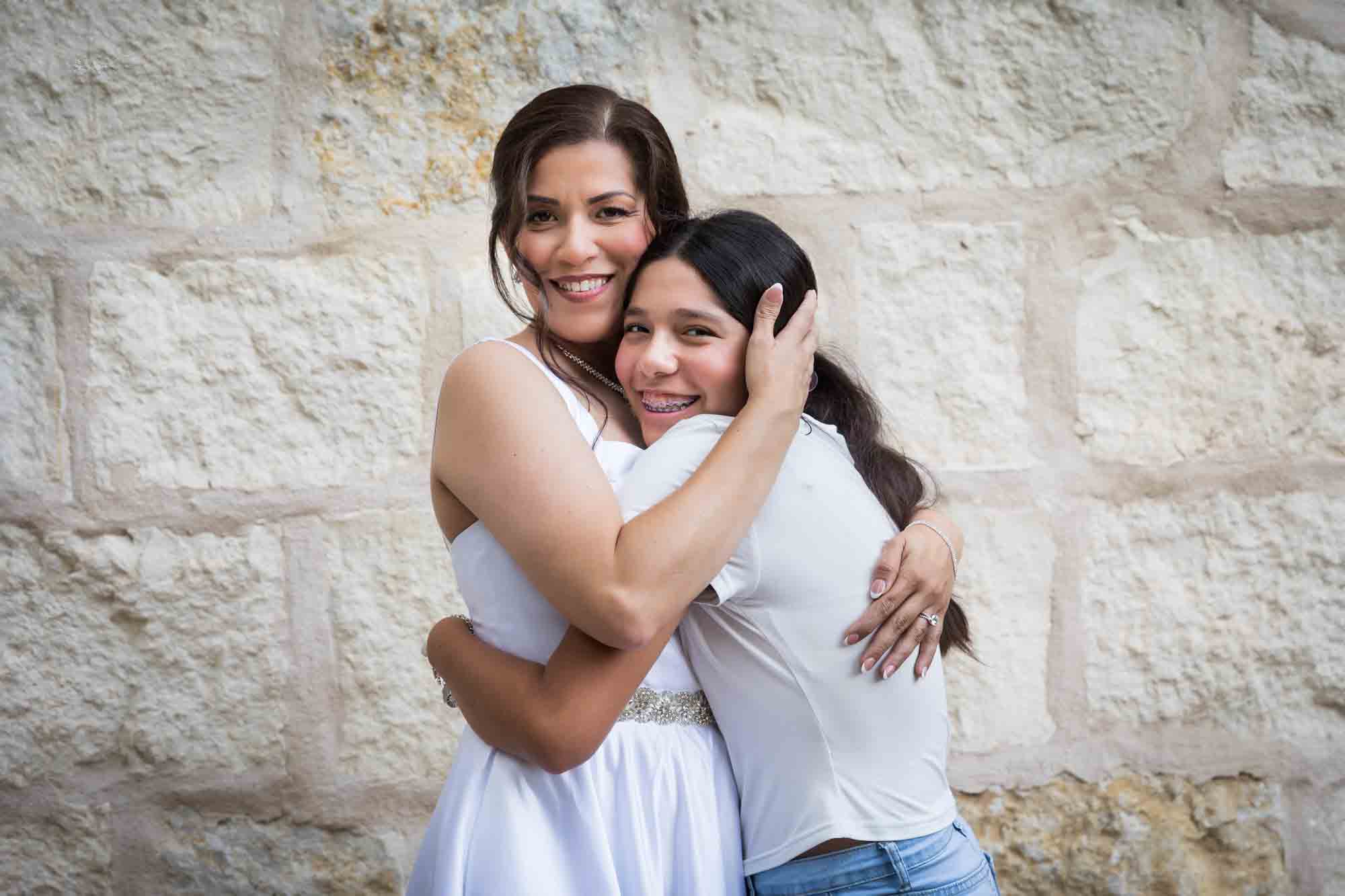 Bride and young girl hugging in front of beige, brick wall during a downtown San Antonio bridal portrait session