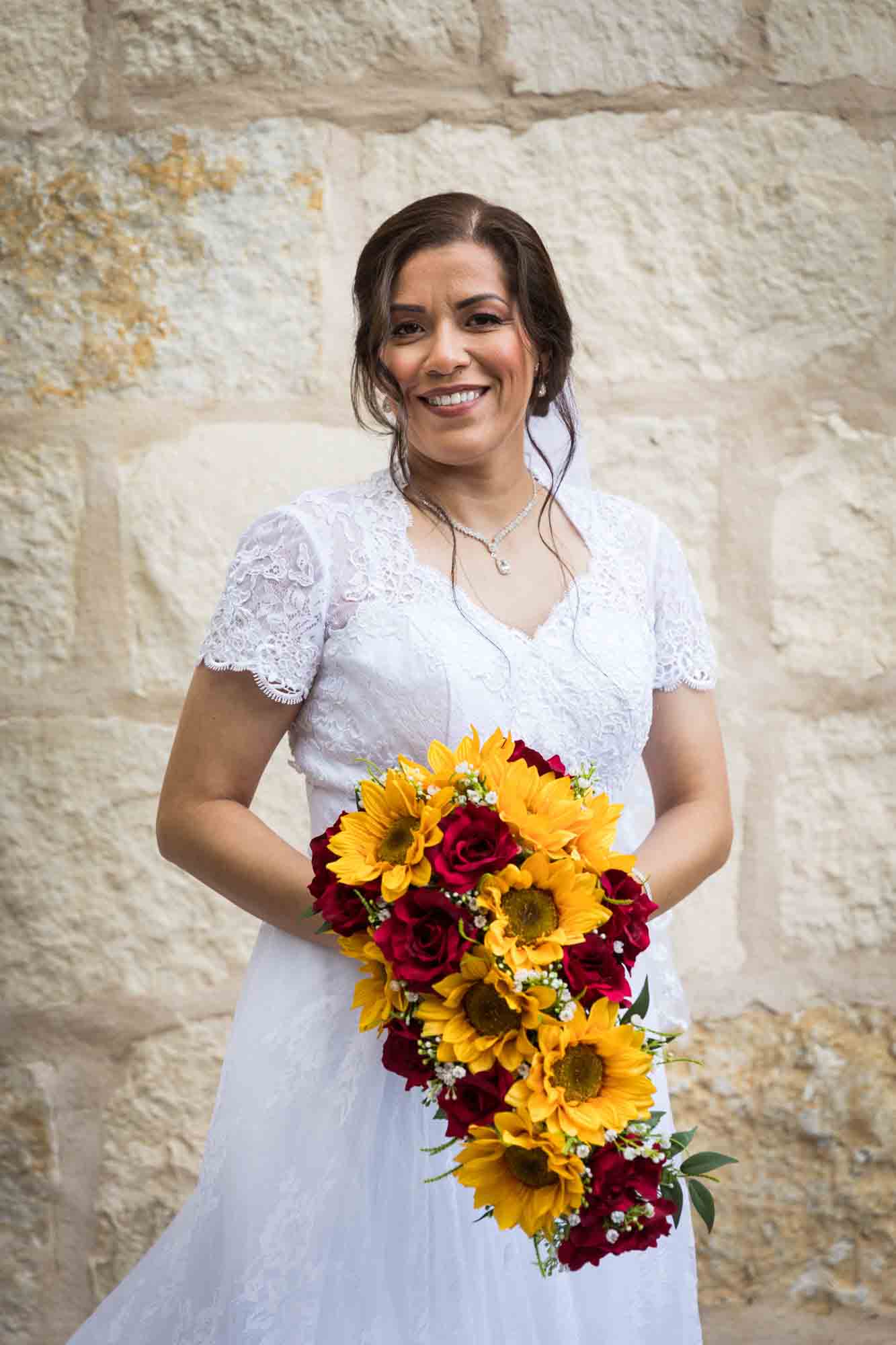 Bride posing in white dress with veil holding flower bouquet in front of beige, brick wall for an article about a downtown San Antonio bridal portrait session