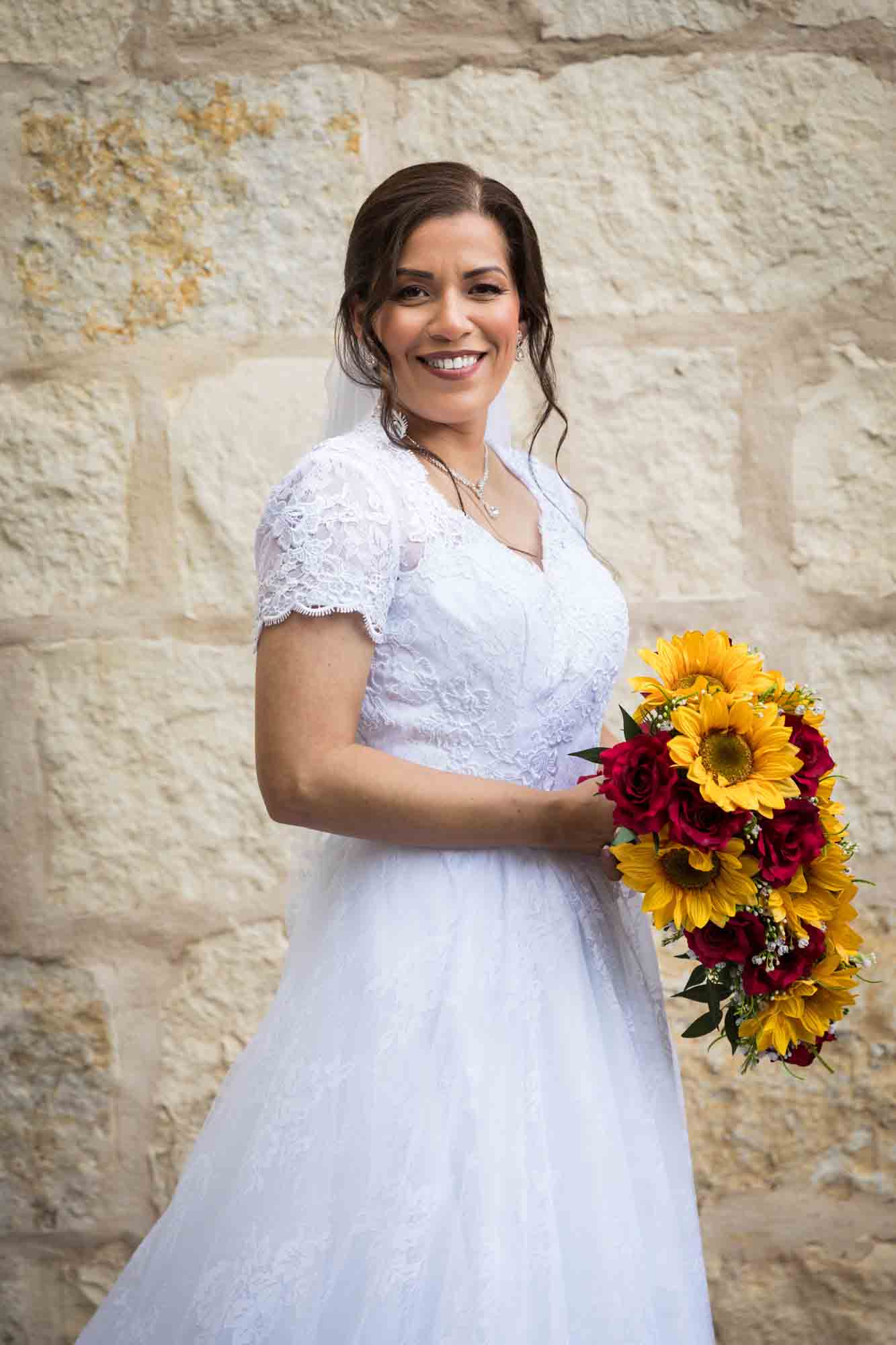 Bride posing in white dress with veil holding flower bouquet in front of beige, brick wall for an article about a downtown San Antonio bridal portrait session