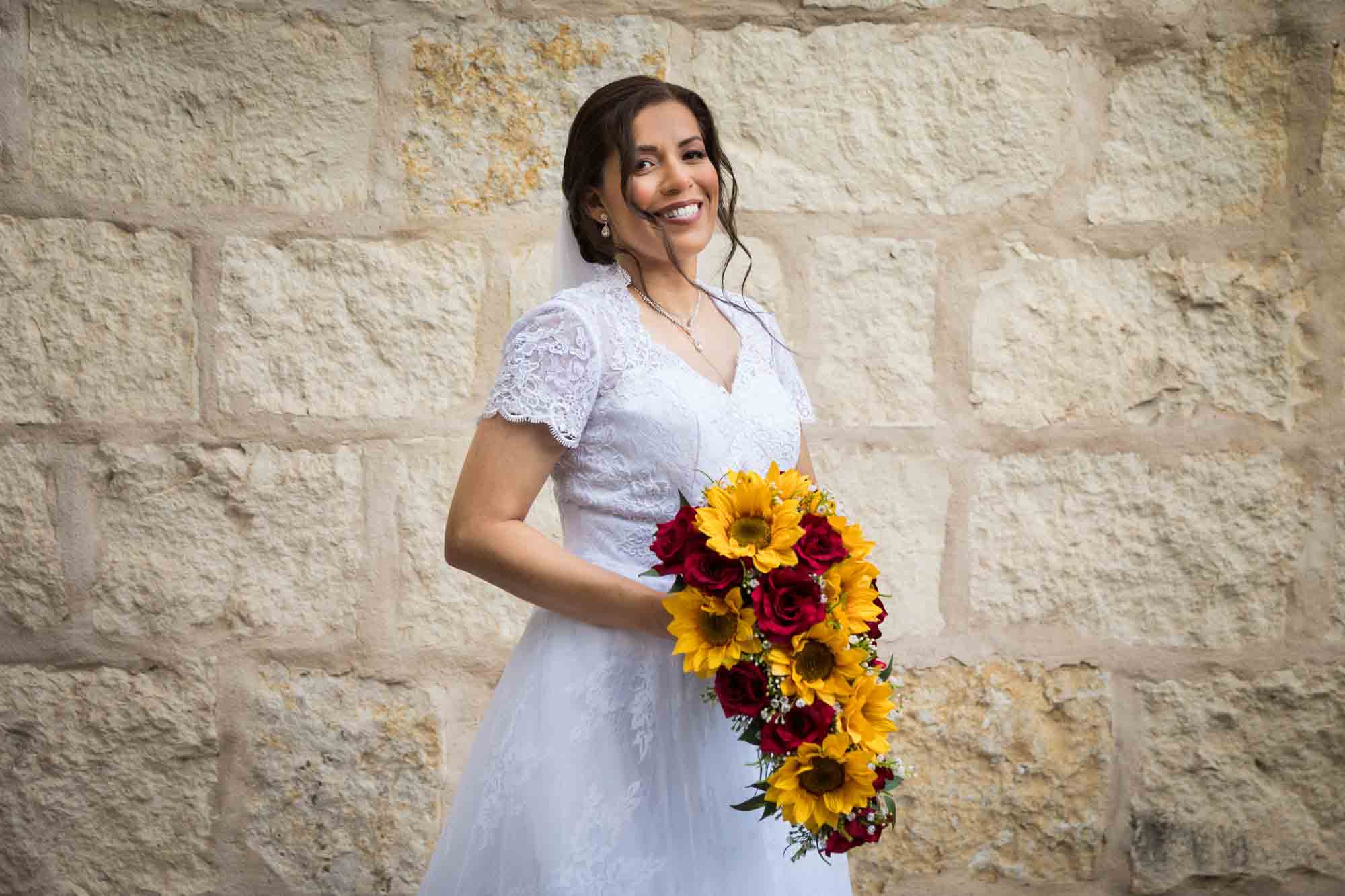 Bride posing in white dress with veil holding flower bouquet in front of beige, brick wall for an article about a downtown San Antonio bridal portrait session