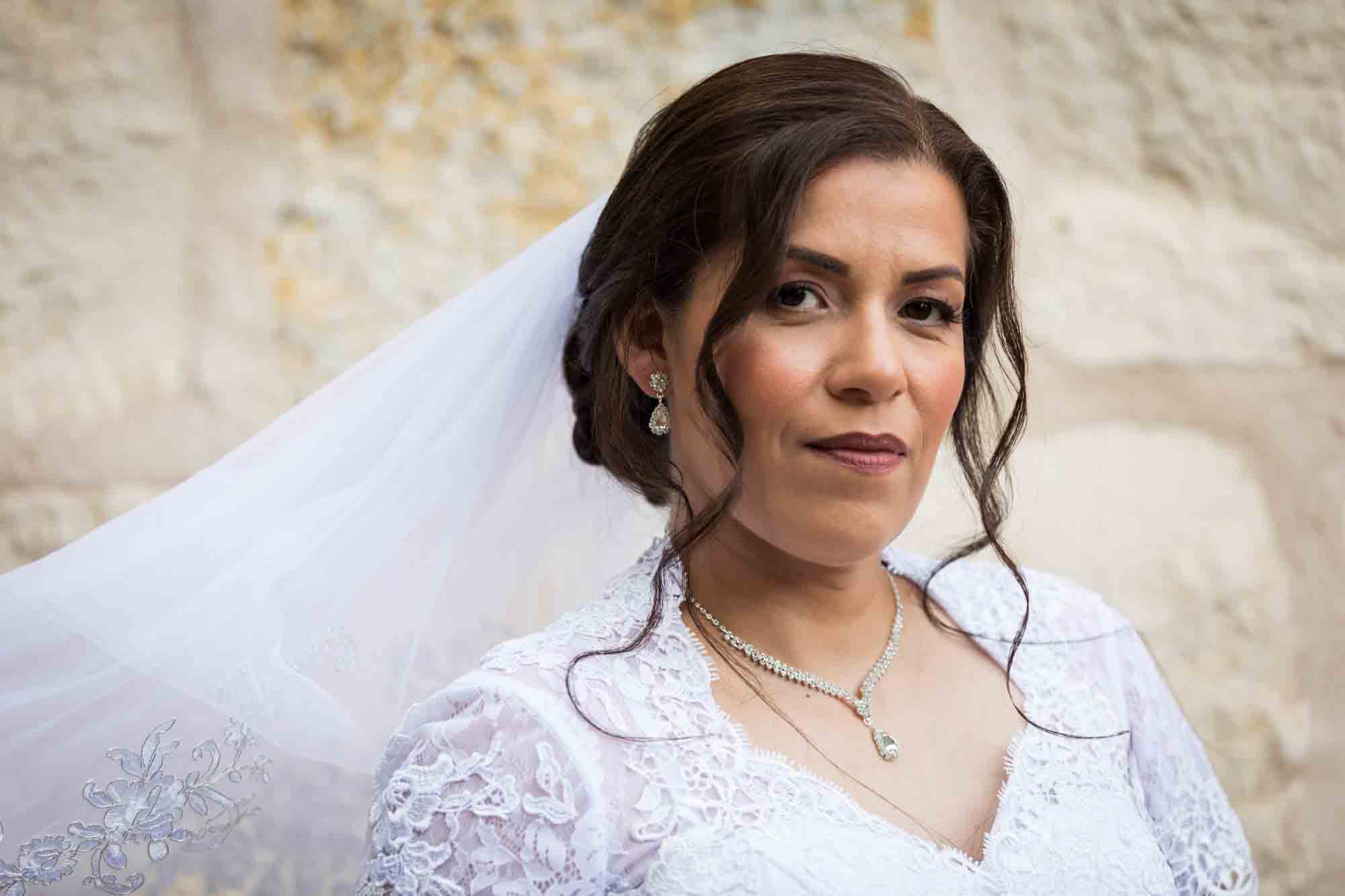 Bride posing in white dress with veil in front of beige, brick wall for an article about a downtown San Antonio bridal portrait session