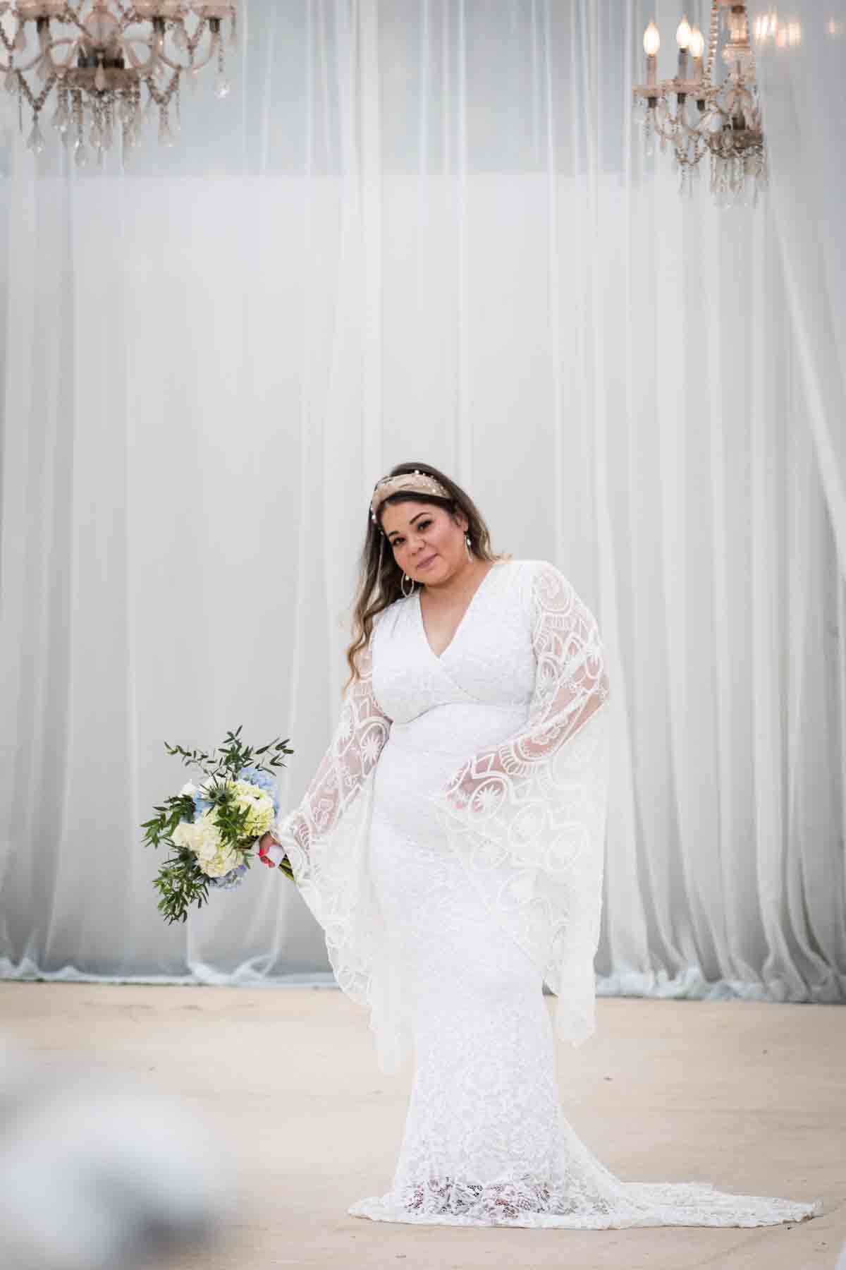 Bride with long sleeves holding a white and blue bridal bouquet in front of long, white curtains and crystal chandeliers