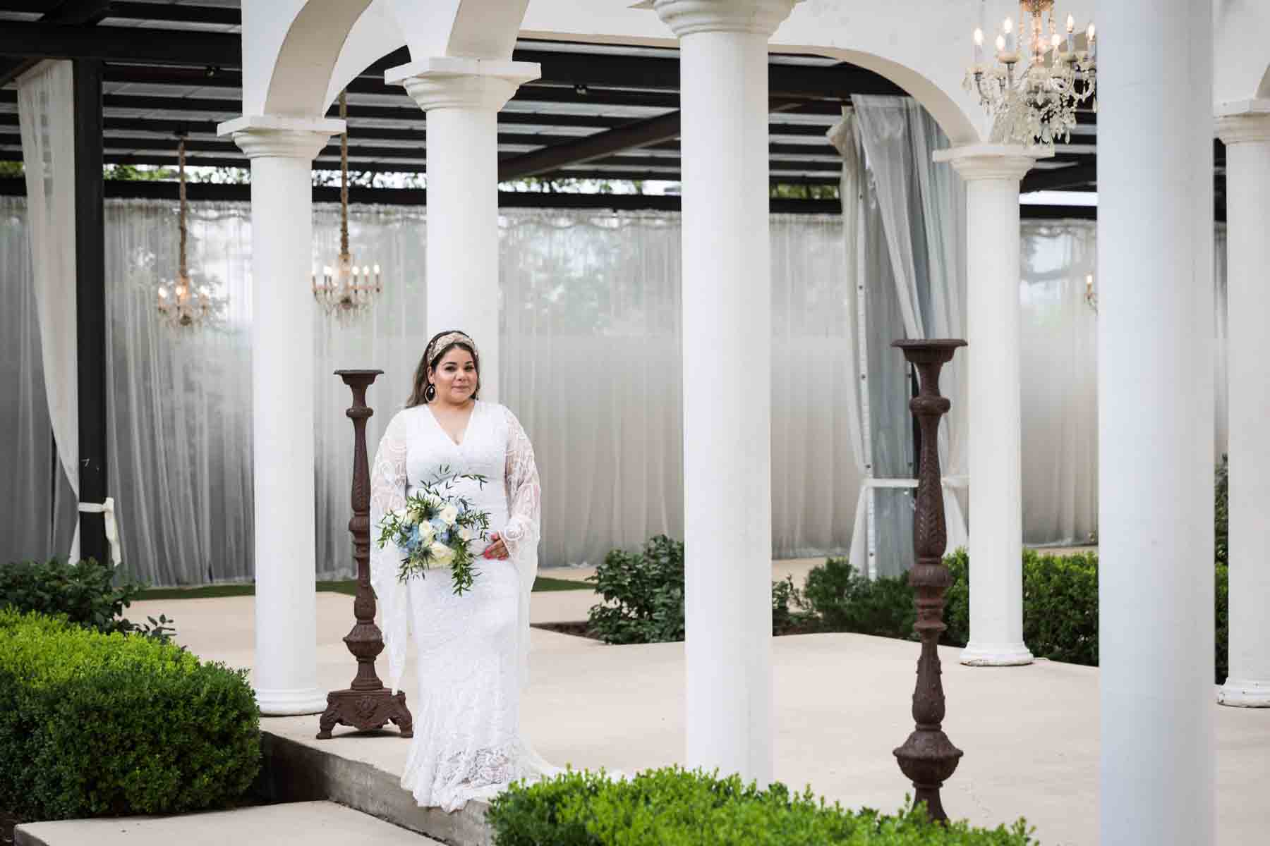 Bride with long sleeves holding a white and blue bridal bouquet in front of white columns and bronze candelabras