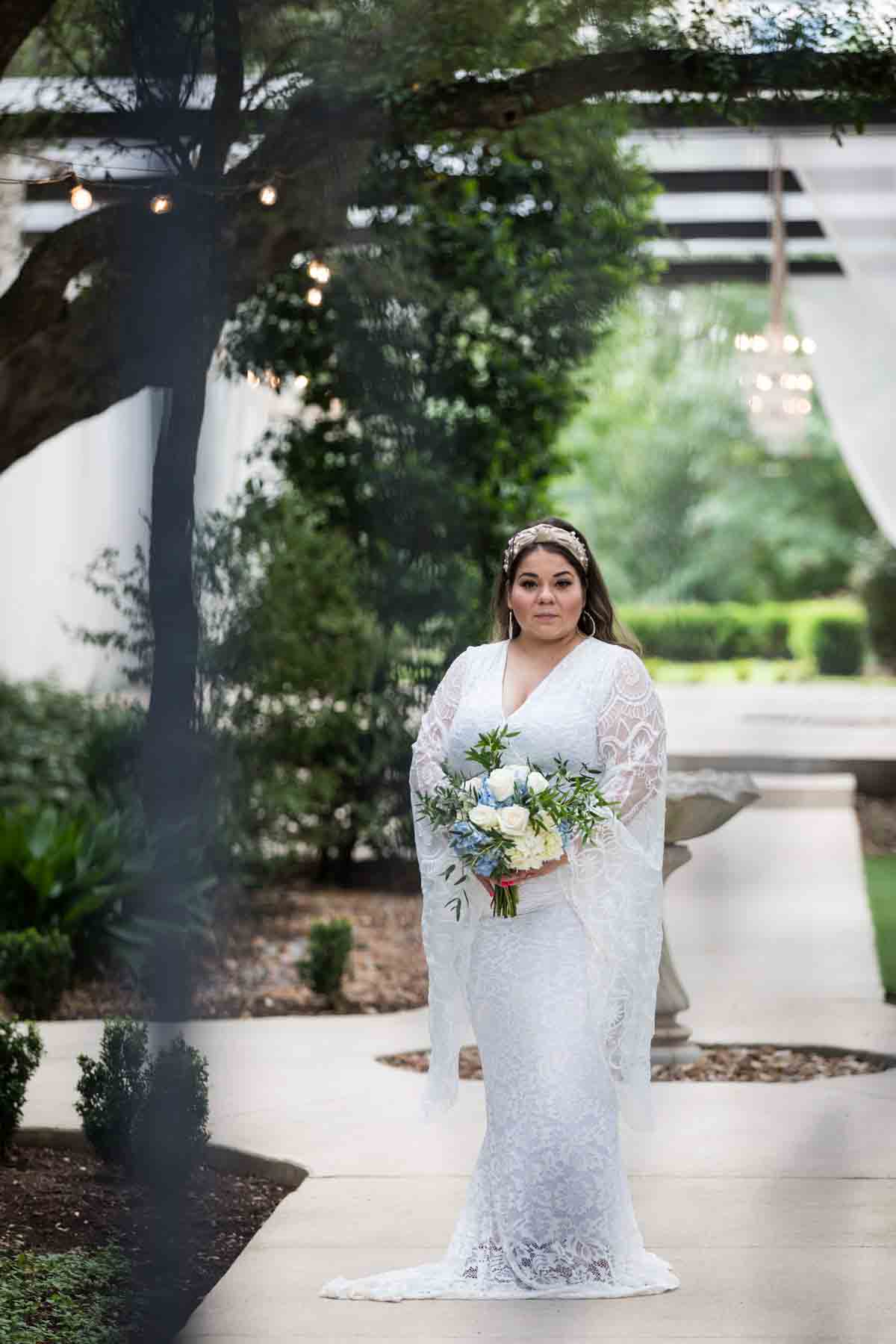 Bride with long sleeves holding a white and blue bridal bouquet on sidewalk as seen through metal gate