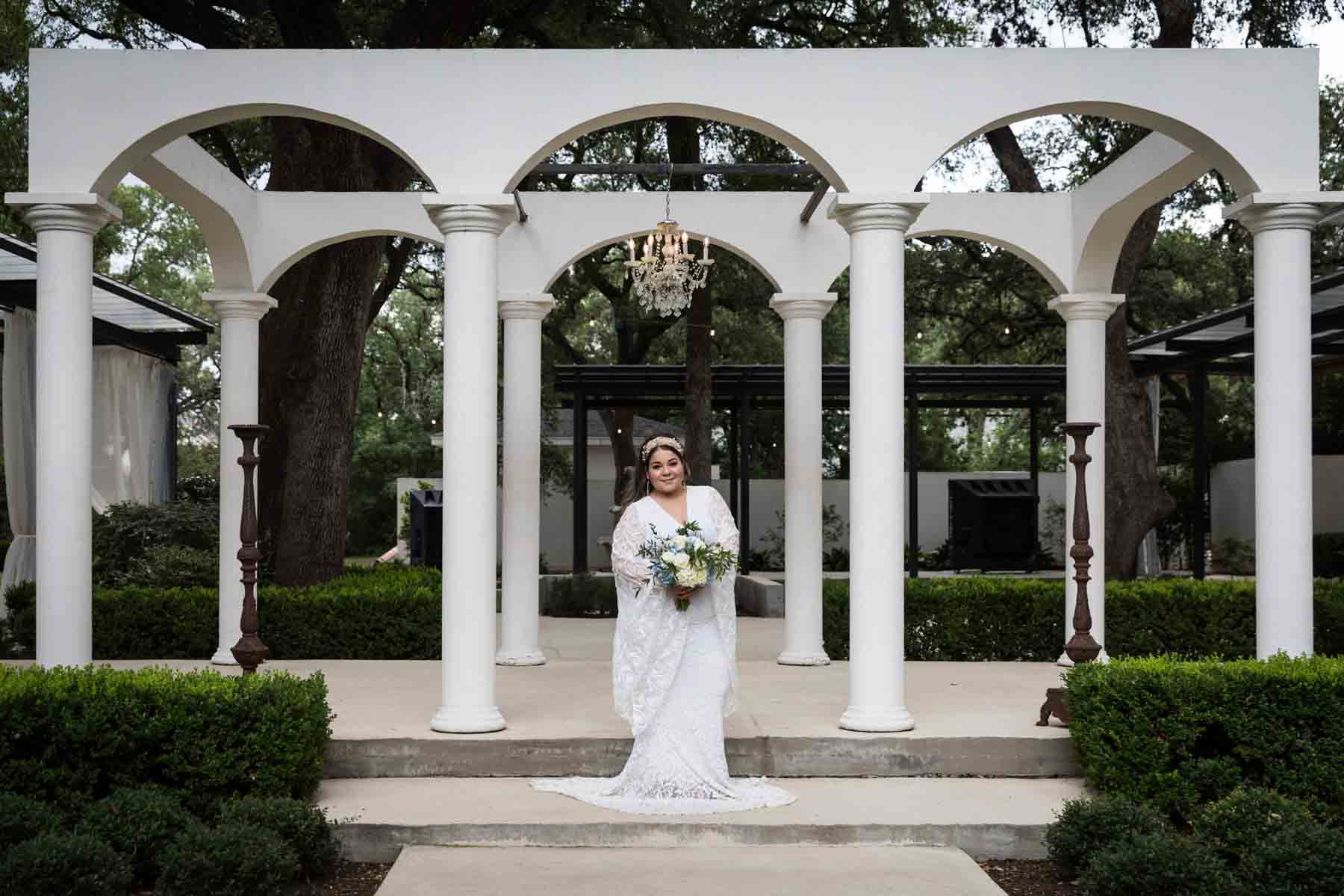 Bride with long sleeves holding a white and blue bridal bouquet in front of long, white colonnade