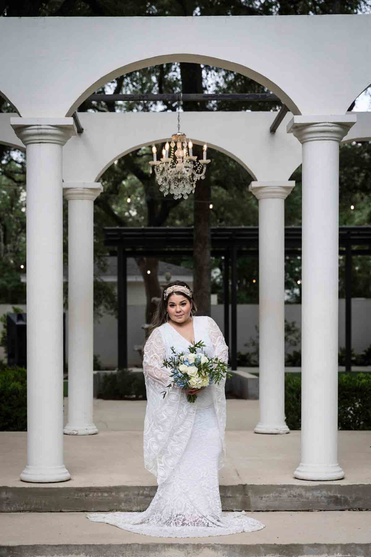 Bride with long sleeves holding a white and blue bridal bouquet in front of long, white colonnade