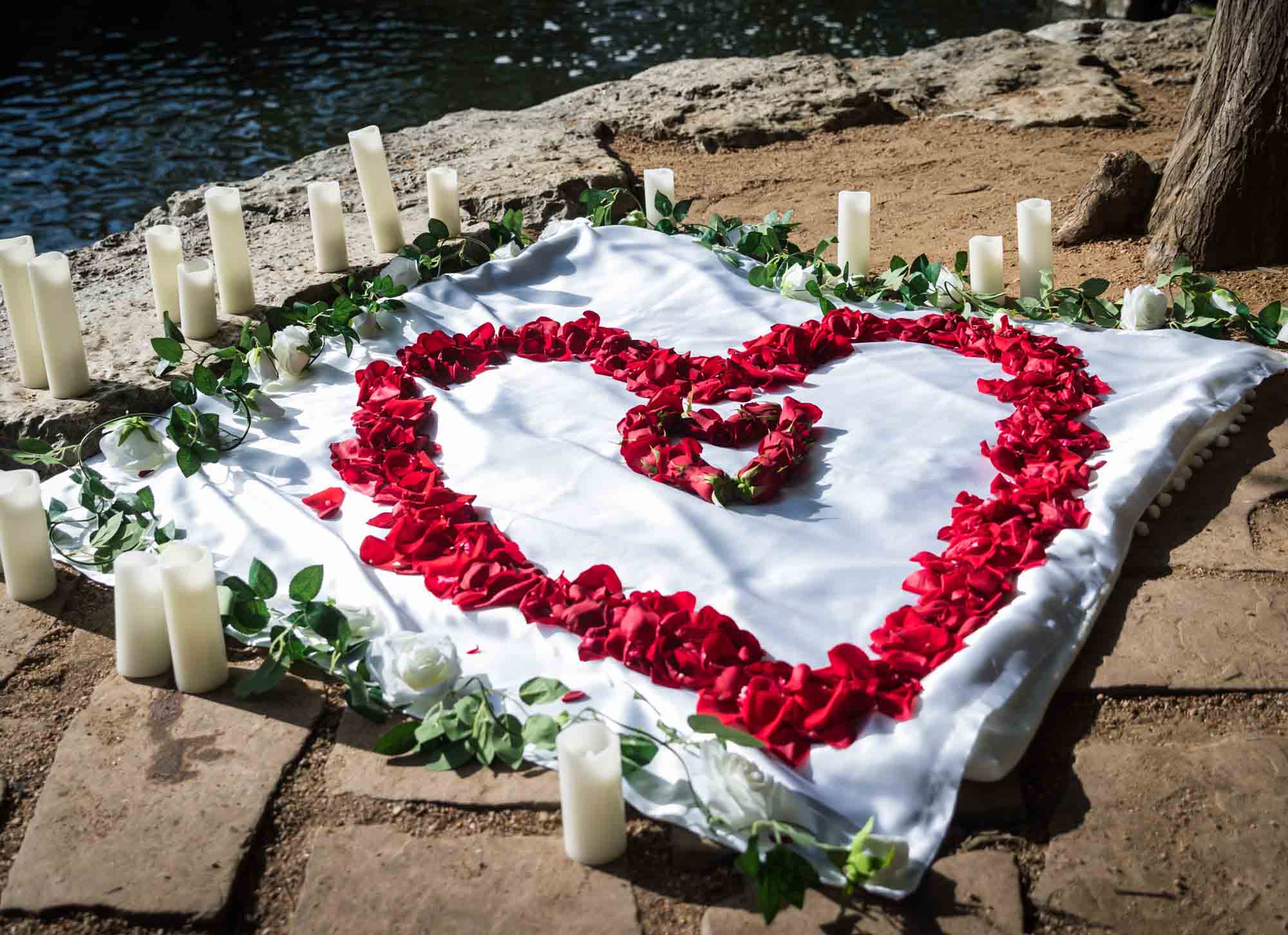 White cloth with red roses scattered in the shape of a heart surrounded by candles on the ground