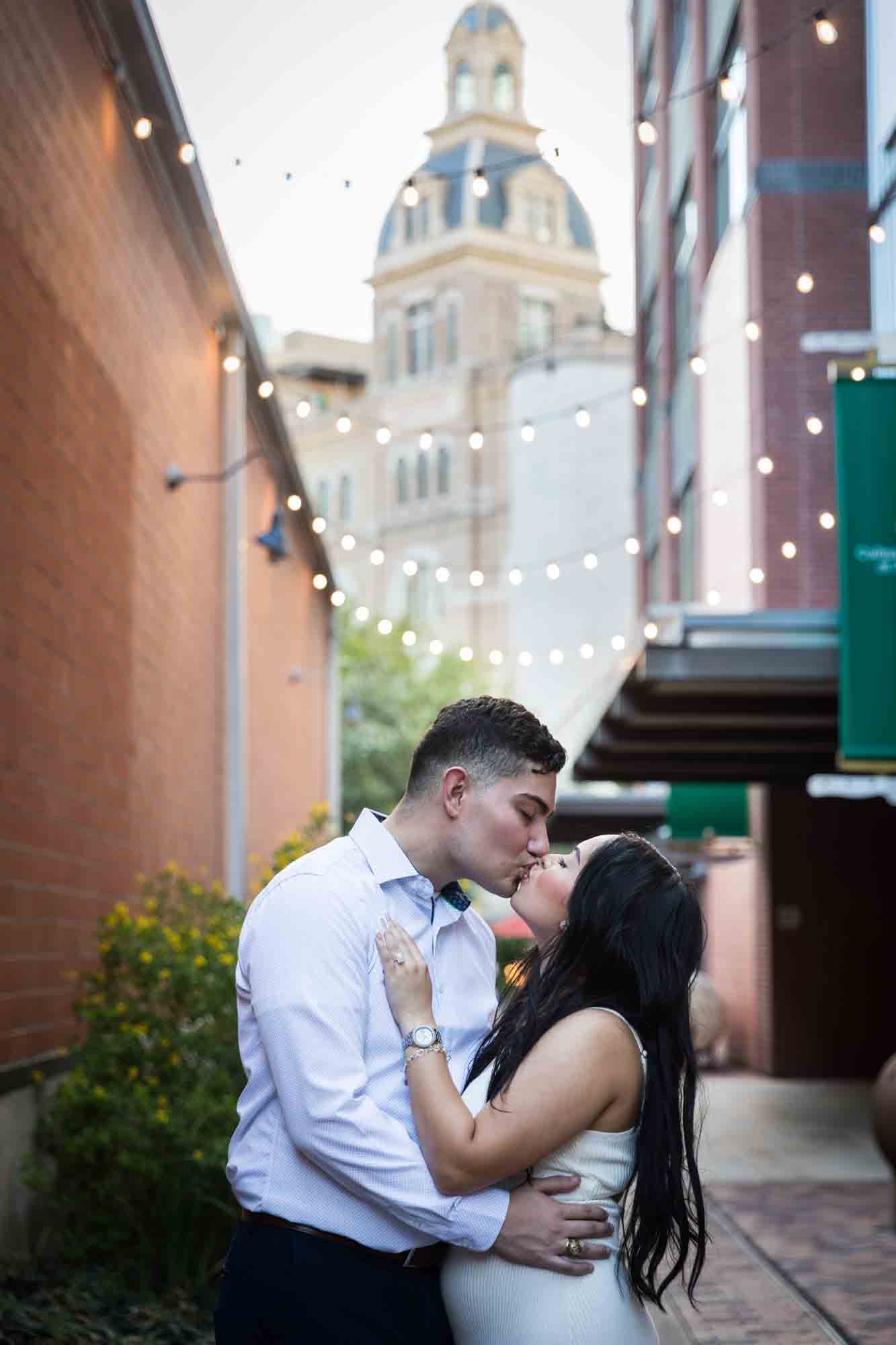 Couple kissing in alleyway with string lights overhead and building in background for an article on Pearl surprise proposal tips