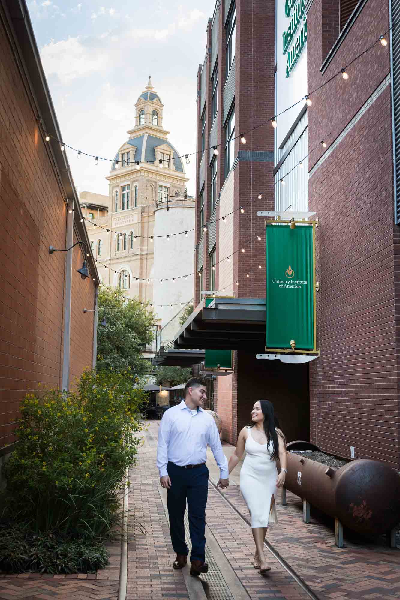 Couple walking down alleyway with string lights overhead and building in background for an article on Pearl surprise proposal tips