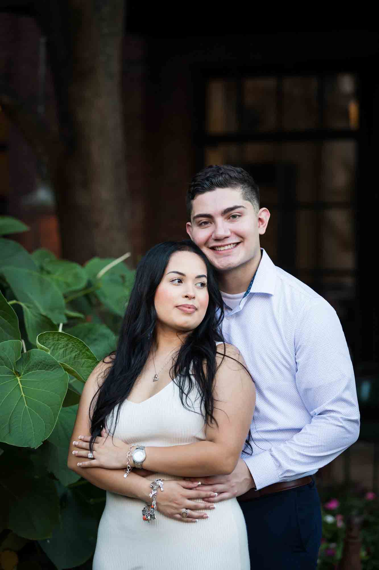 Couple hugging in front of large green leaves for an article on Pearl surprise proposal tips