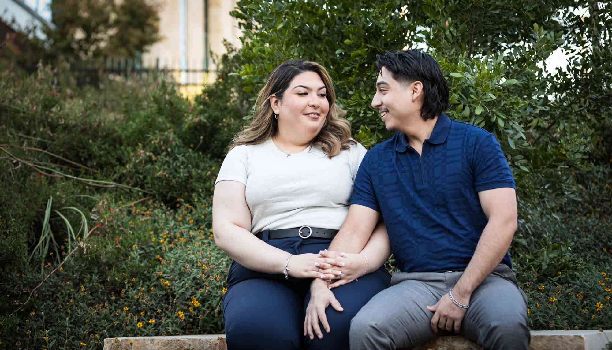 Couple sitting on bench in front of green bushes during a downtown San Antonio photo shoot