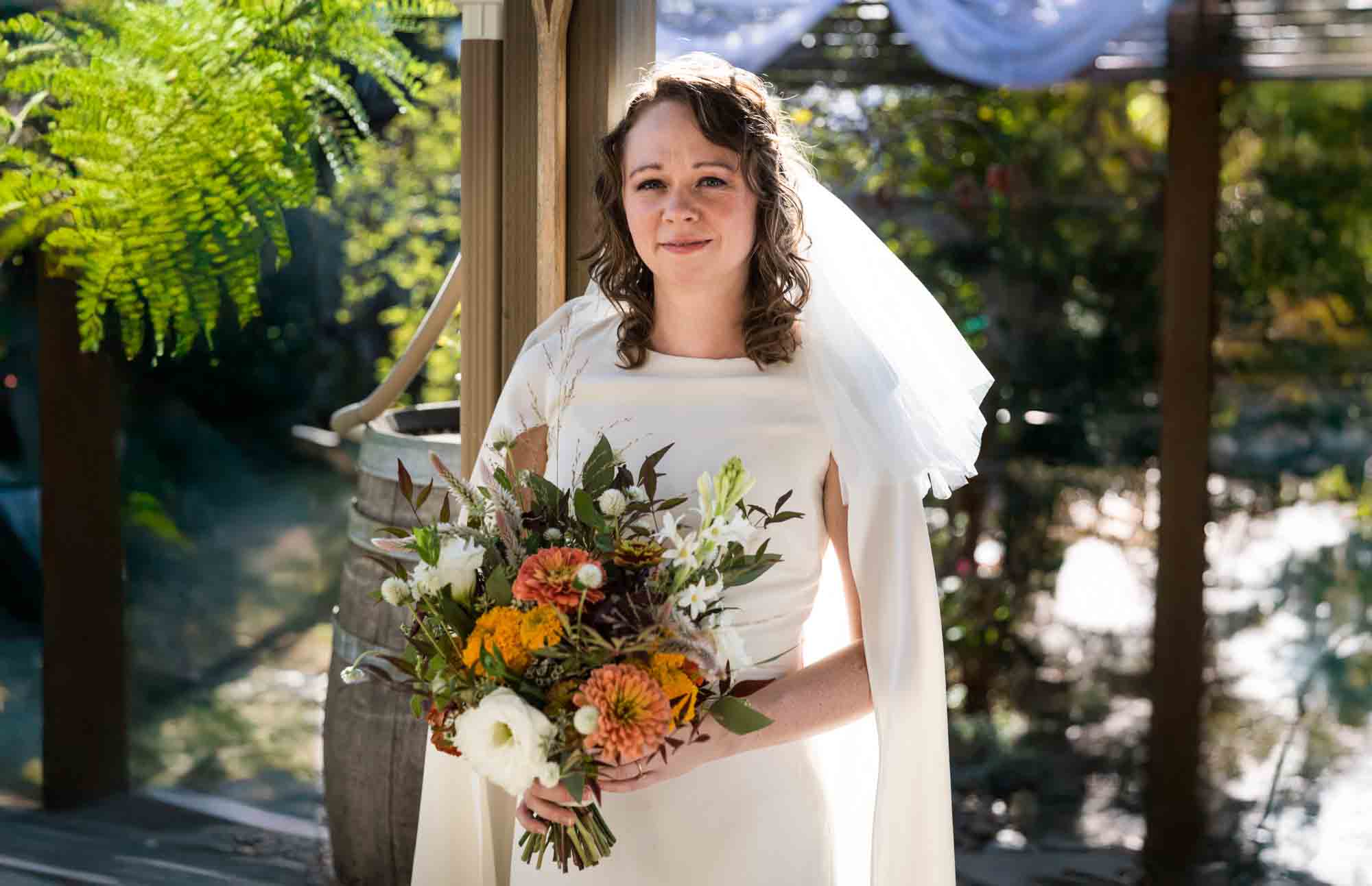 Bride wearing white dress and veil and holding flower bouquet in front of post at a Boerne backyard wedding