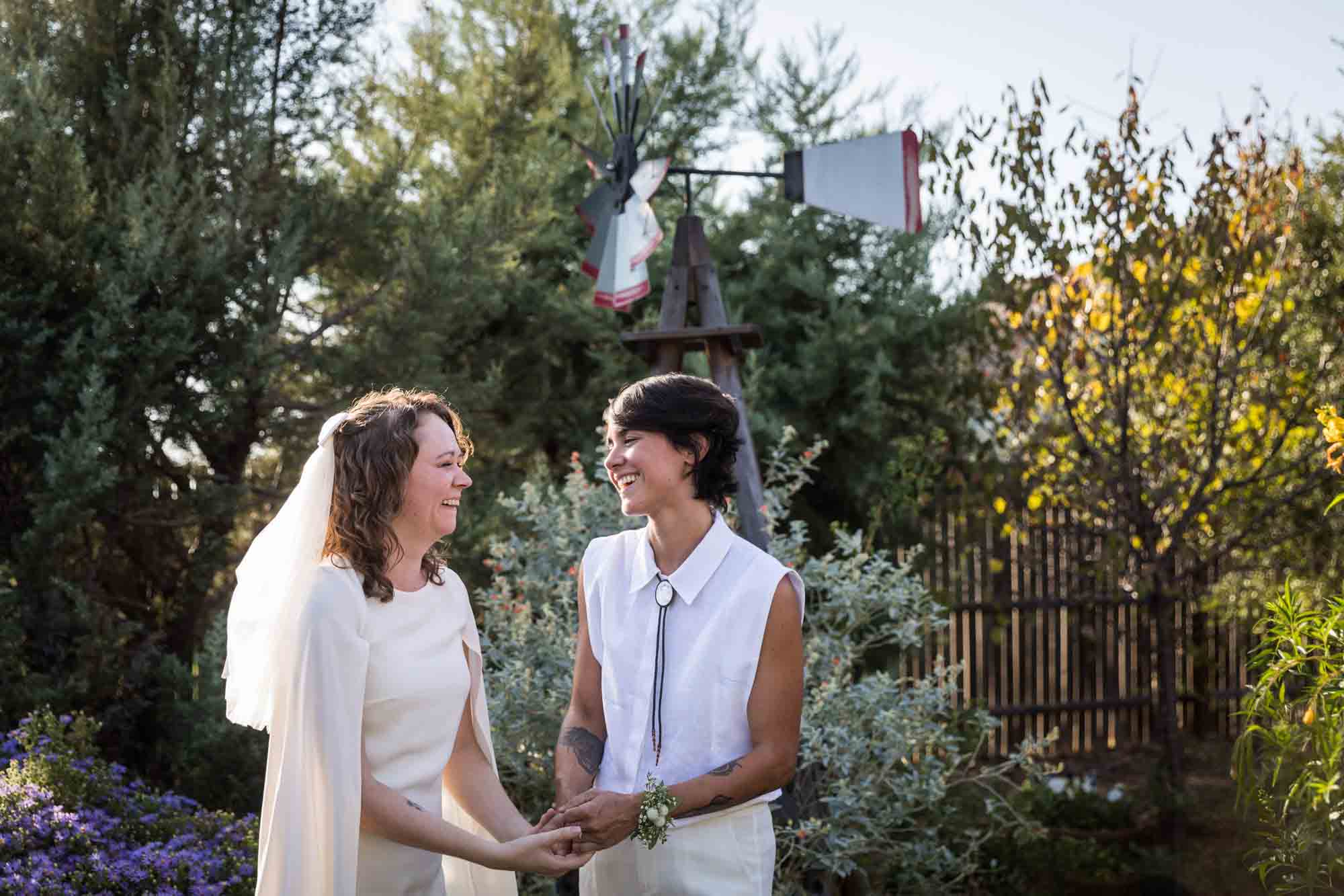 Two brides wearing white and holding hands surrounded by plants at a Boerne backyard wedding