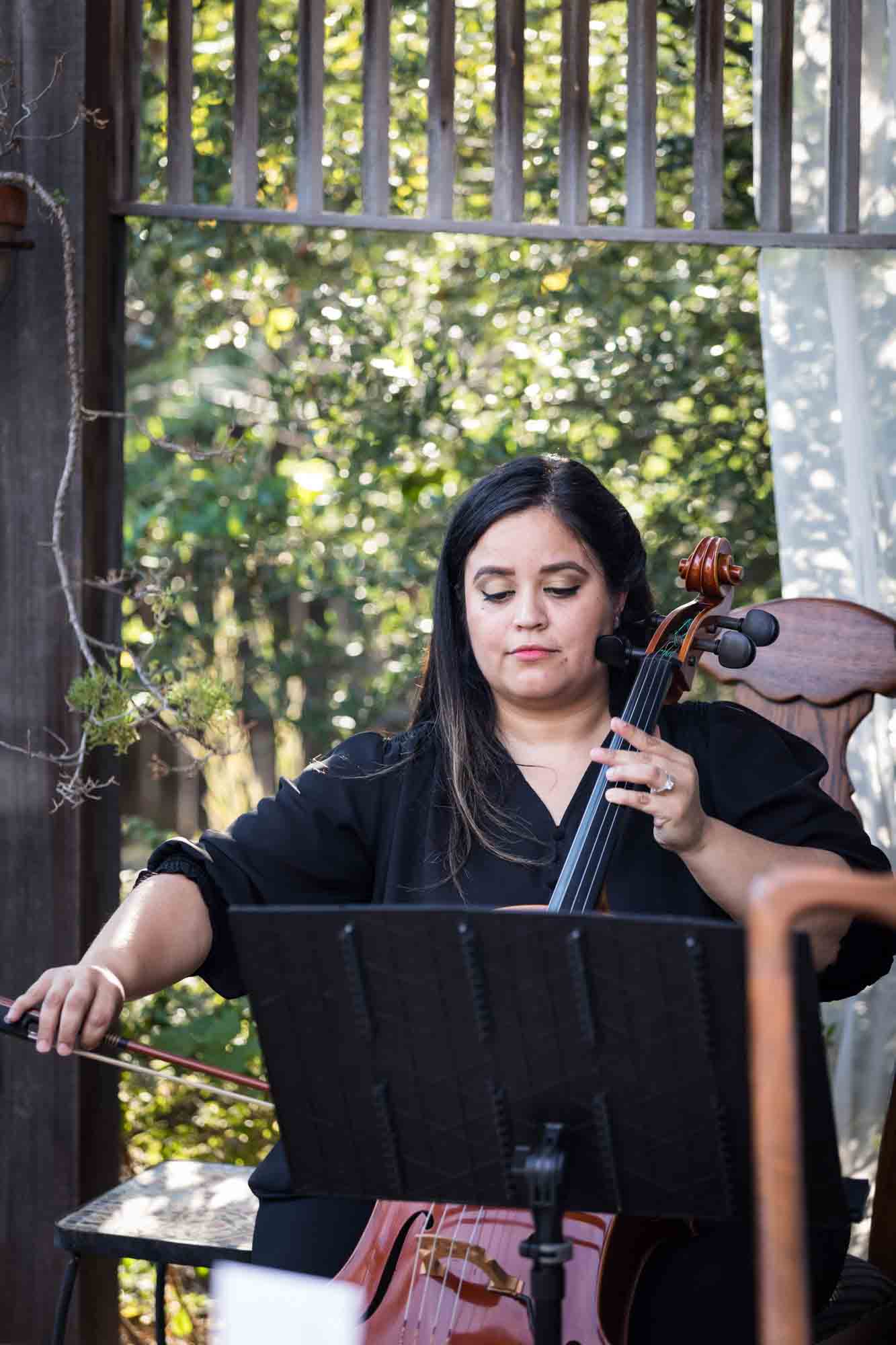 Female cellist wearing black and playing cello in front of plants