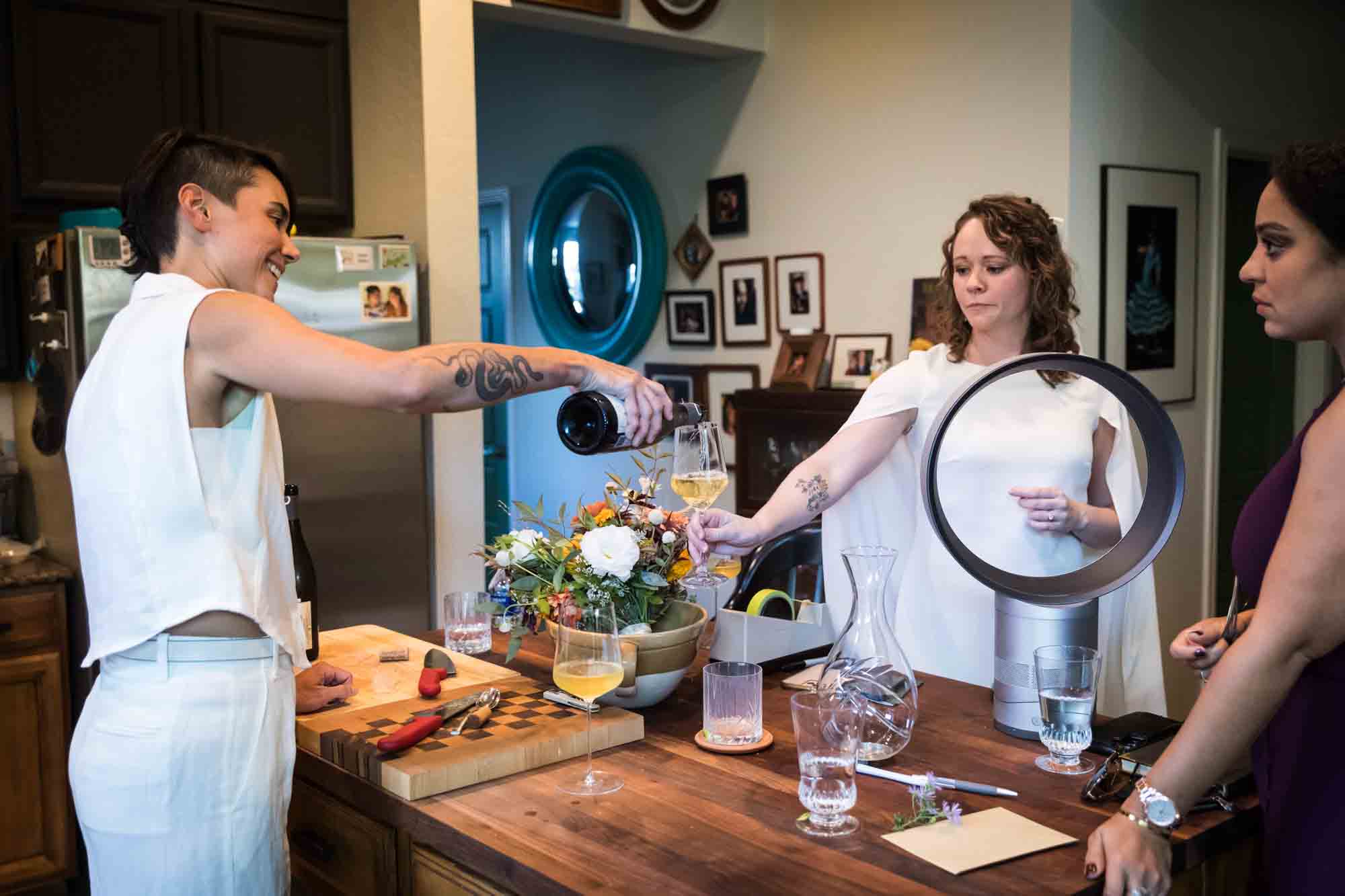 Bride pouring wine in glass for other bride in kitchen