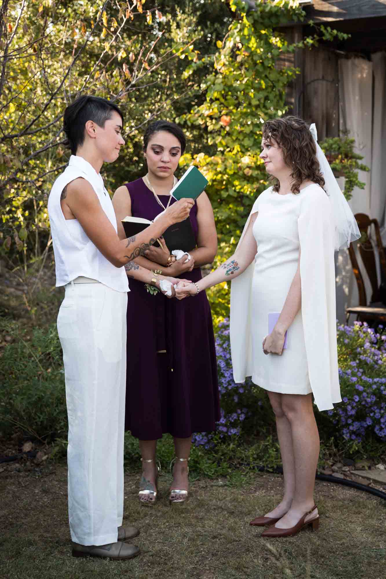 Two brides holding hands and exchanging vows in front of officiant and guests at a Boerne backyard wedding