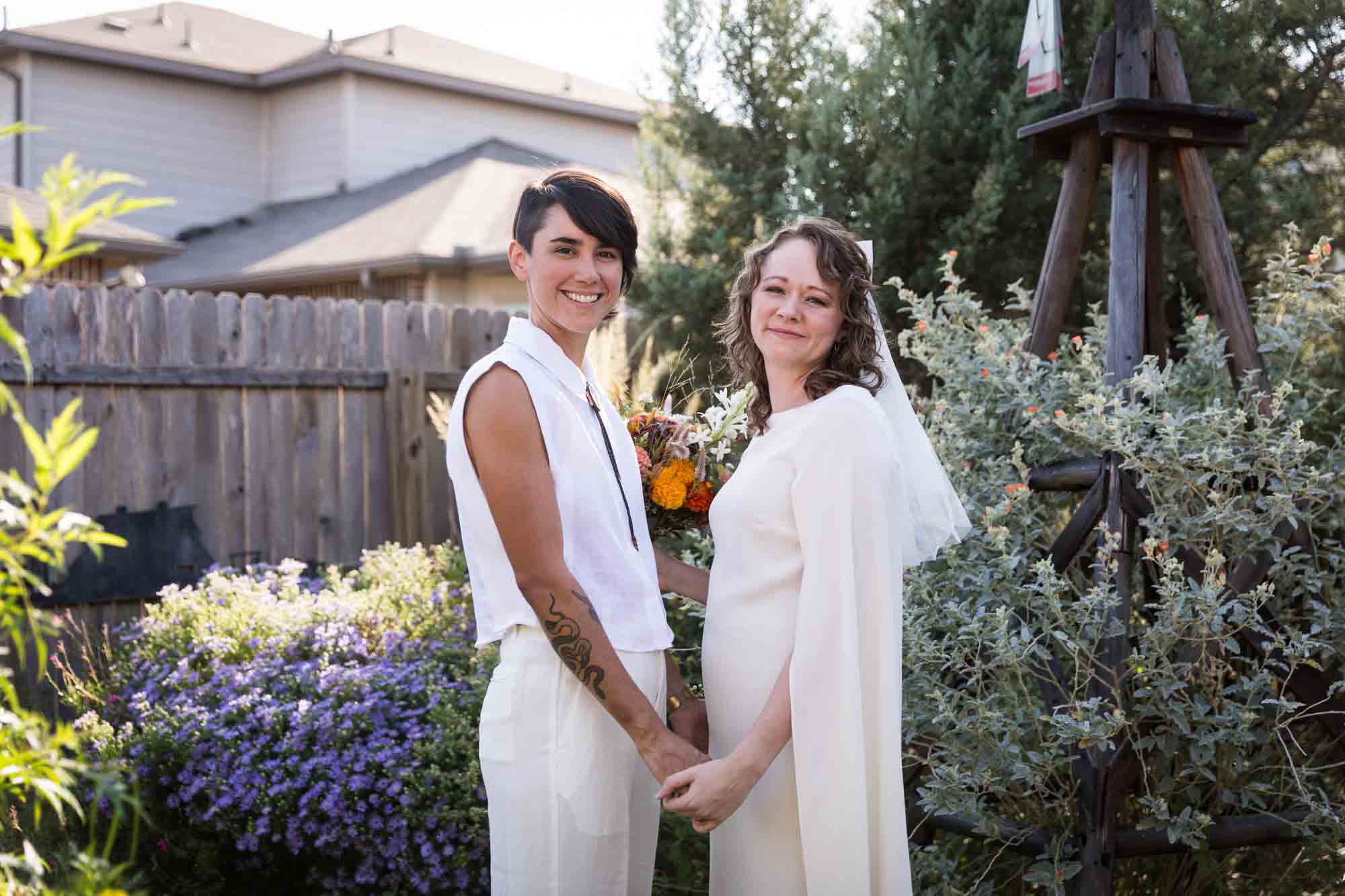 Two brides wearing white and holding hands surrounded by plants at a Boerne backyard wedding