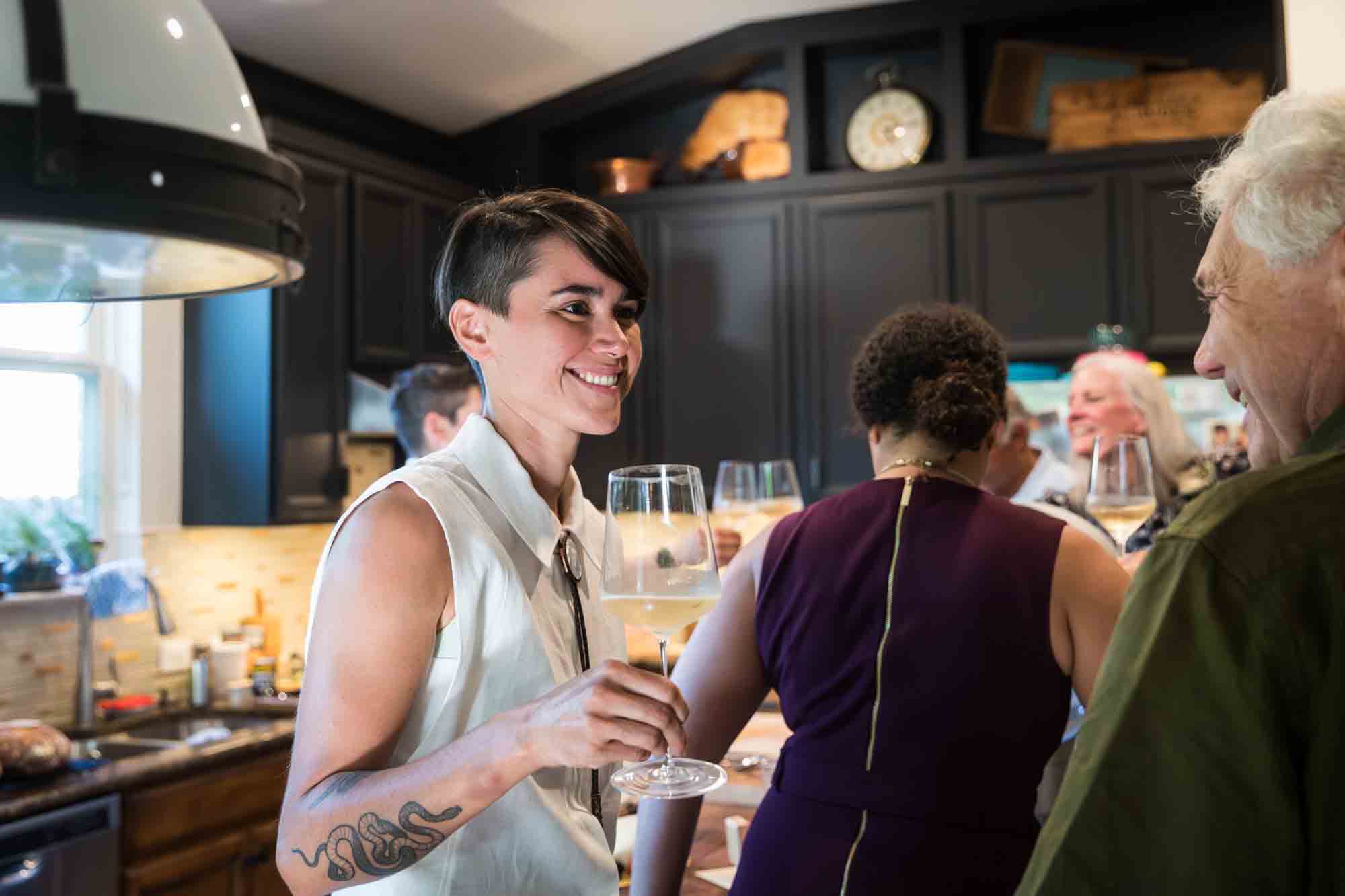 Bride wearing sleeveless white shirt and holding wine glass in kitchen