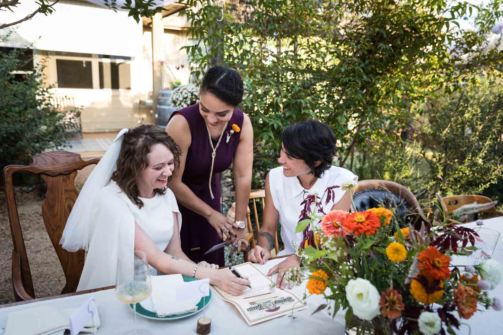 Two brides seated at table signing marriage license with officiant at a Boerne backyard wedding