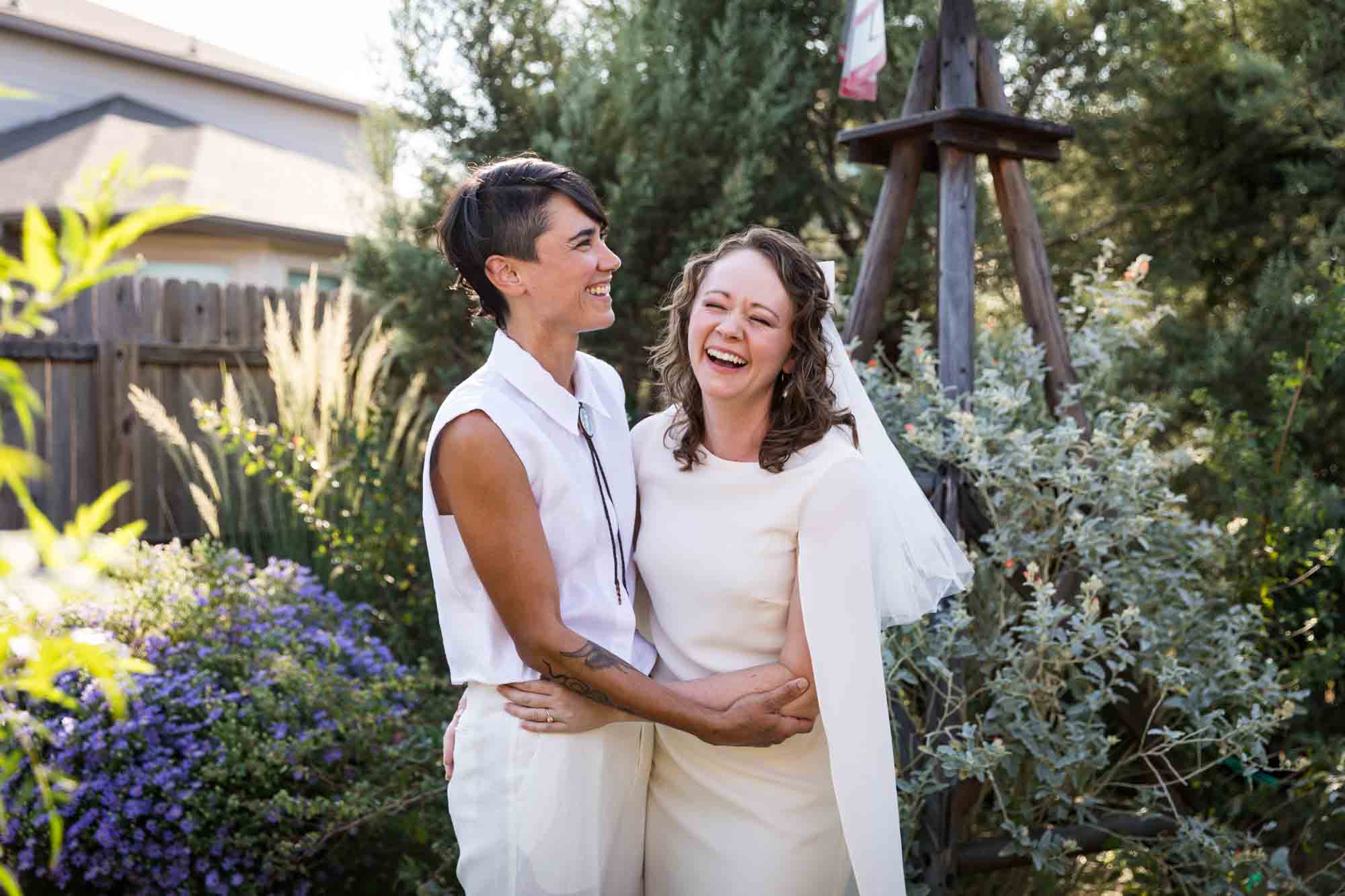 Two brides wearing white and hugging surrounded by plants at a Boerne backyard wedding